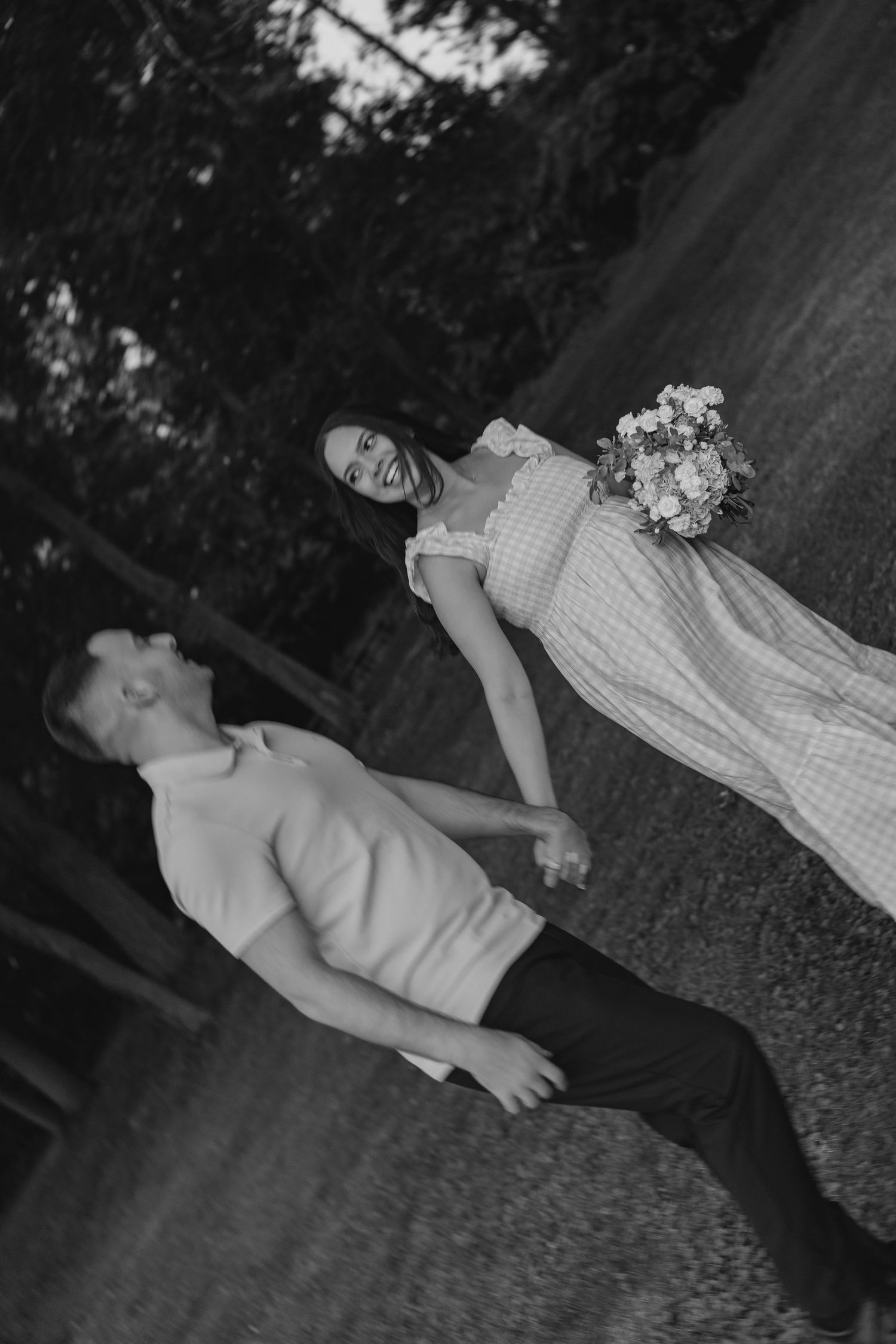 Black and white photo of smiling pregnant woman holding bouquet leading partner by hand along a tree-lined path.