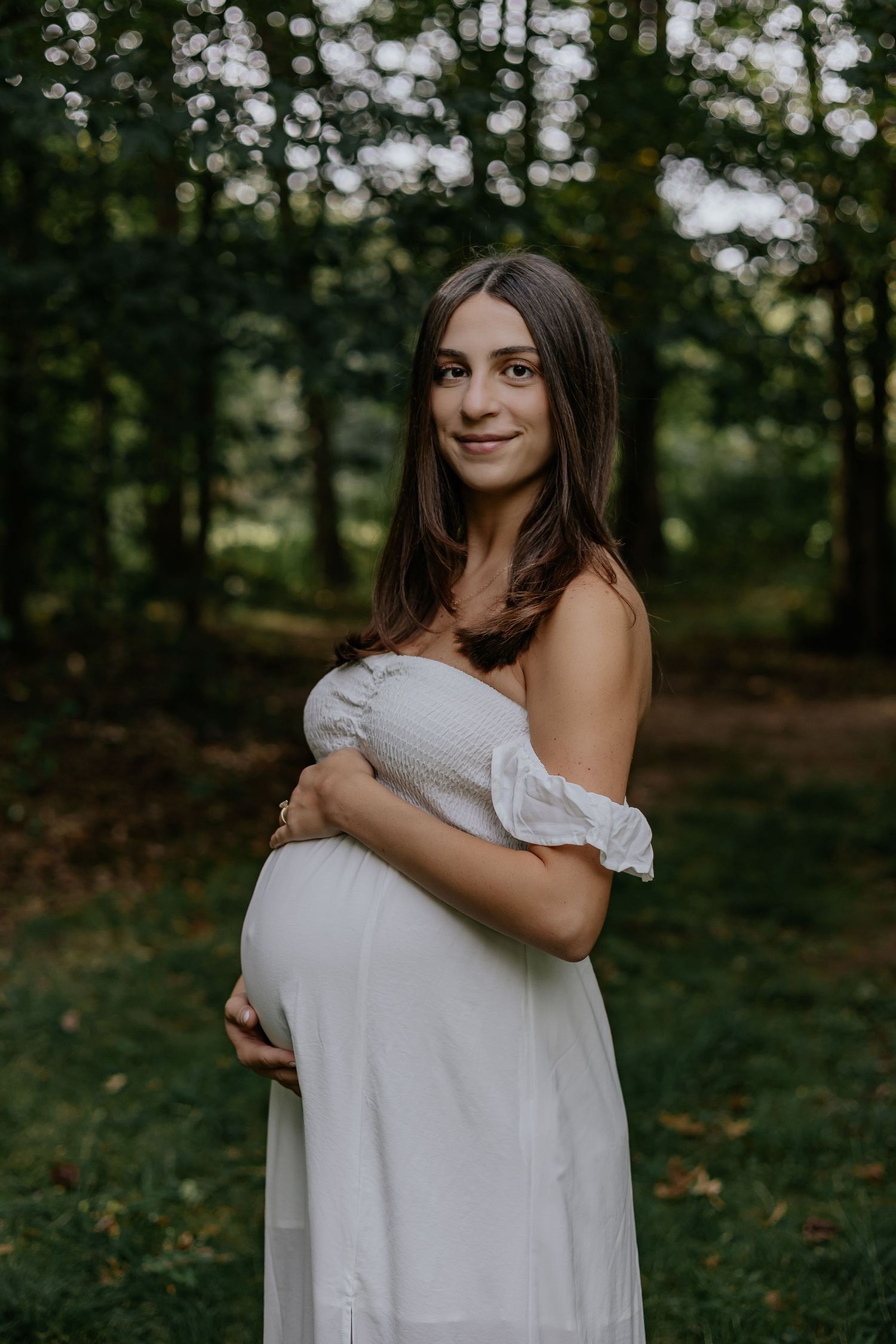 Pregnant woman in white off-shoulder dress cradling baby bump in lush green forest setting.
