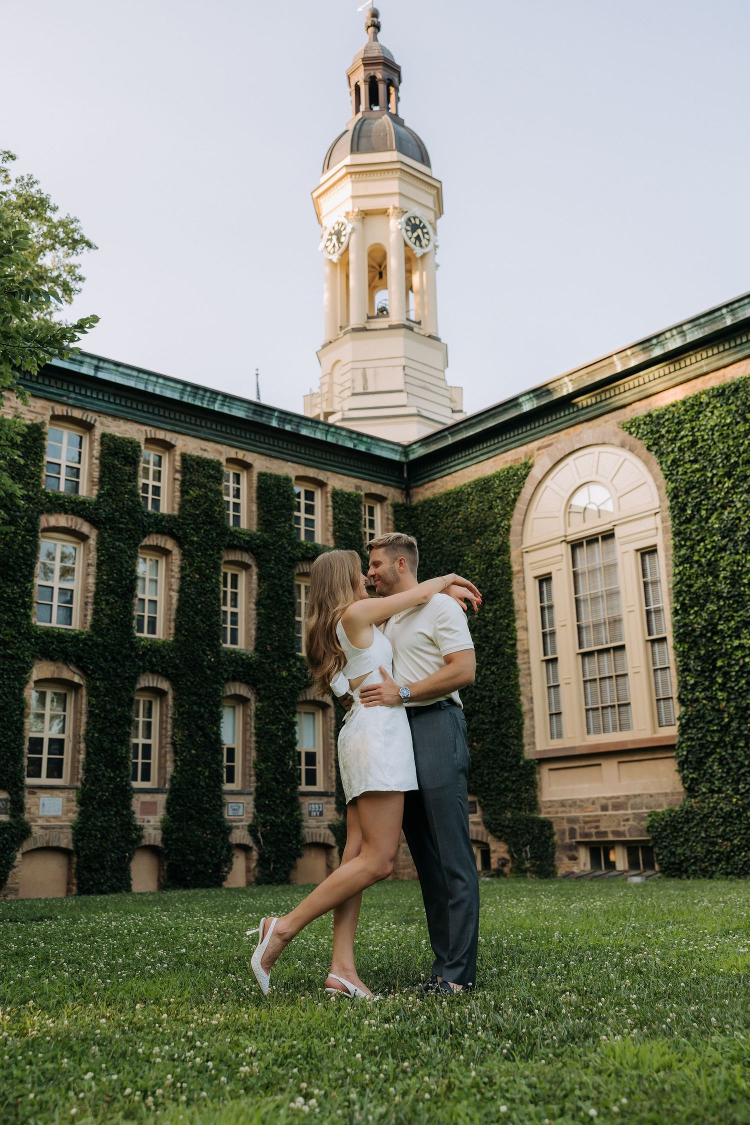 Couple embracing and dancing on green lawn in front of ivy-covered building with a clock tower at golden hour.