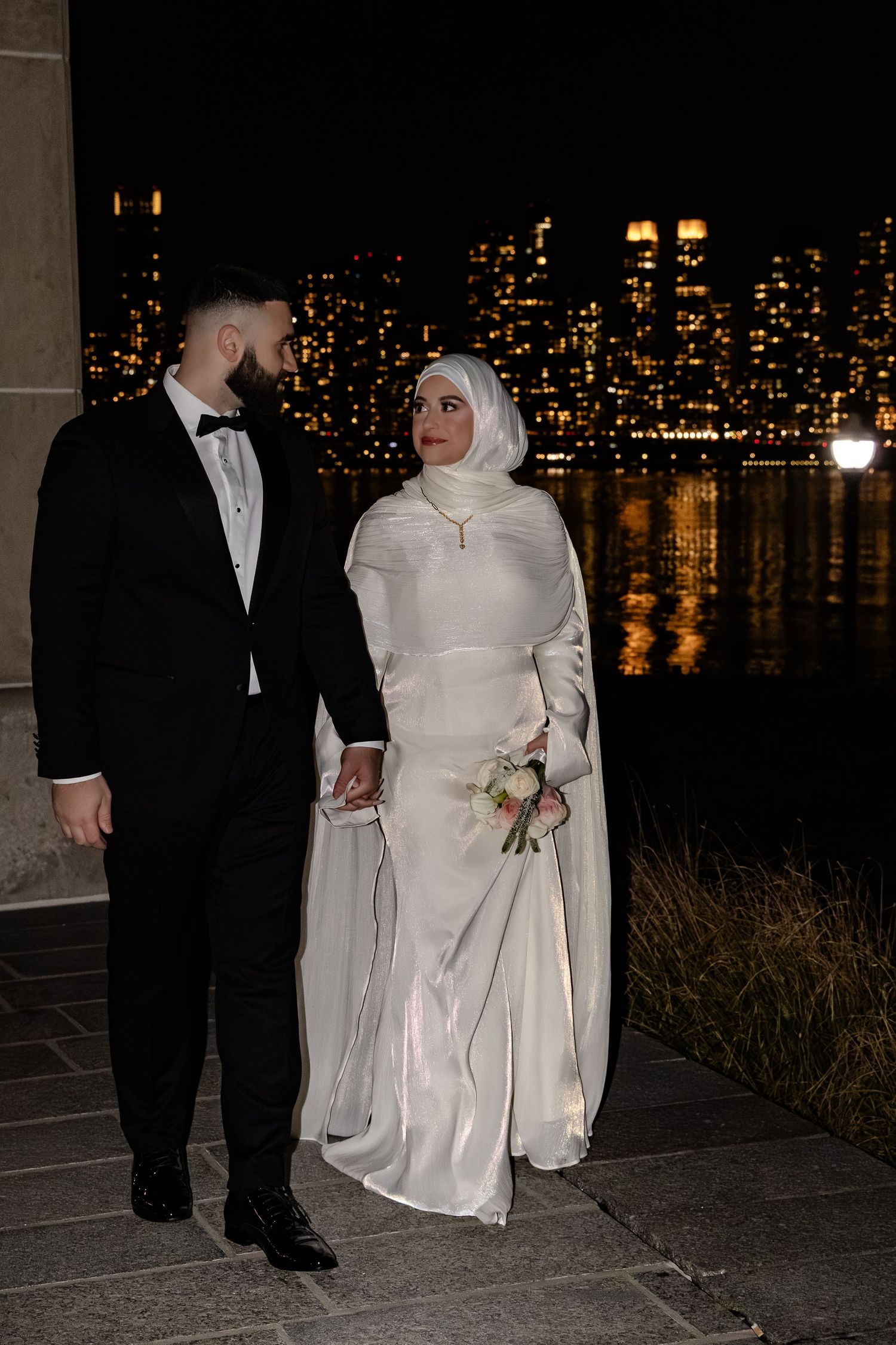 Muslim bride and groom holding hands walking by waterfront with glowing city skyline at night.