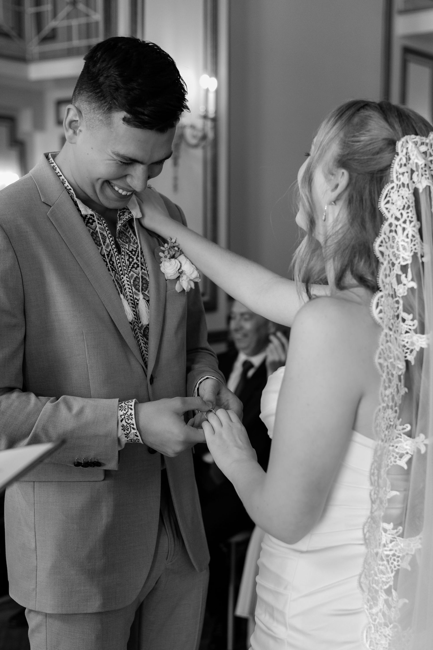 Smiling groom in gray suit receives wedding ring from bride in lace veil, black and white.