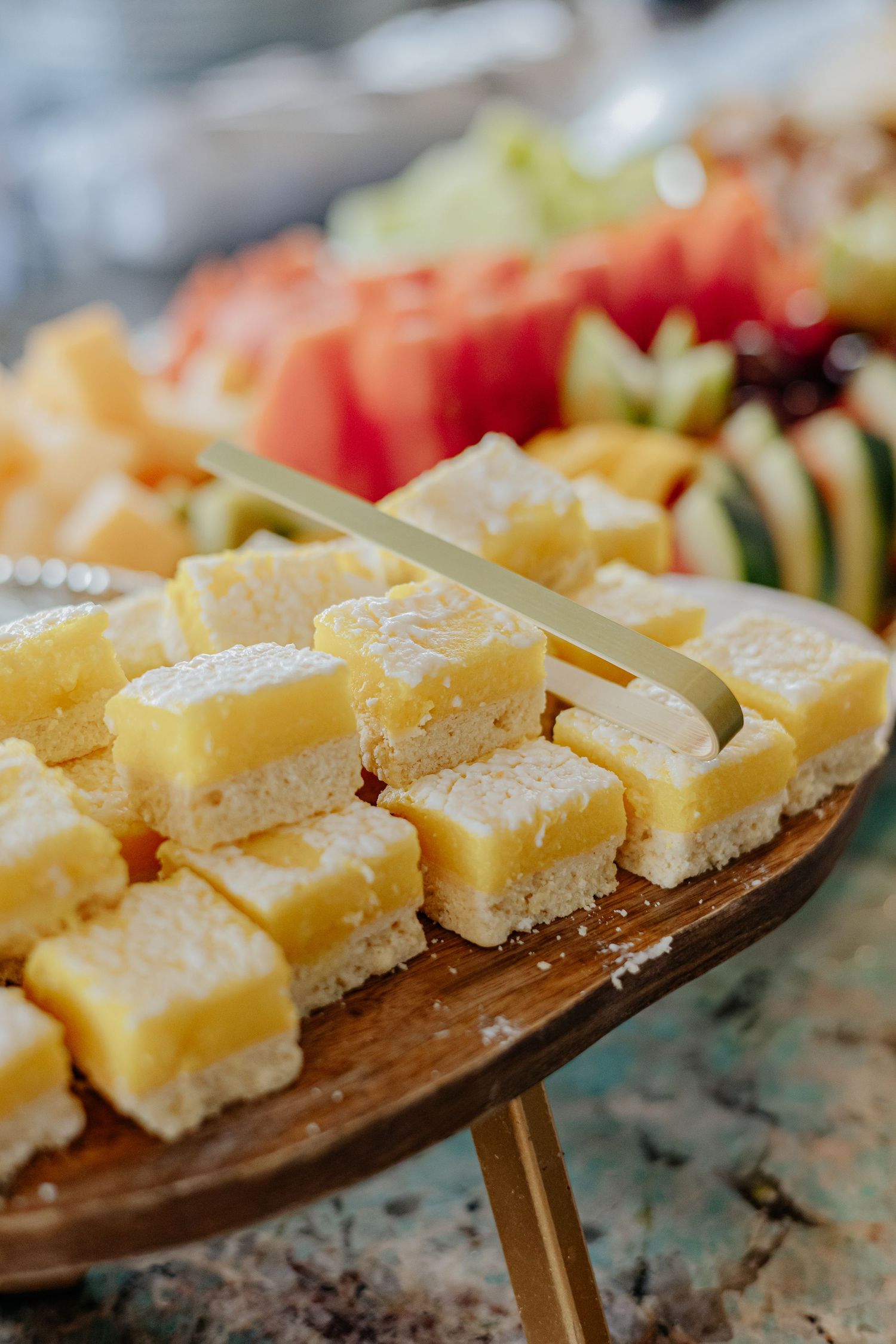 Lemon bar squares on a wooden stand with fresh fruit platter in the background.