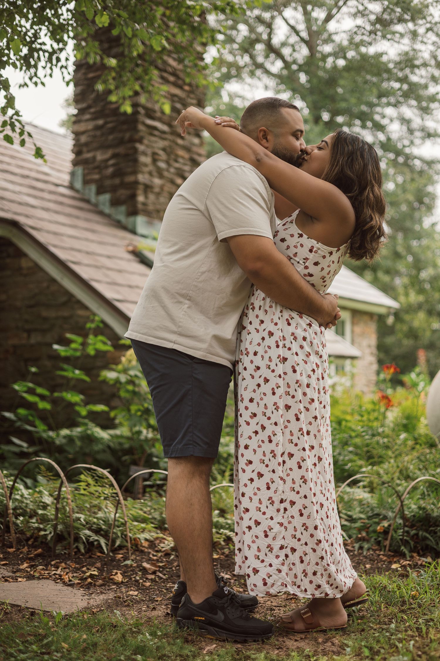 Couple sharing a romantic kiss while embracing in a lush garden beside a stone cottage.