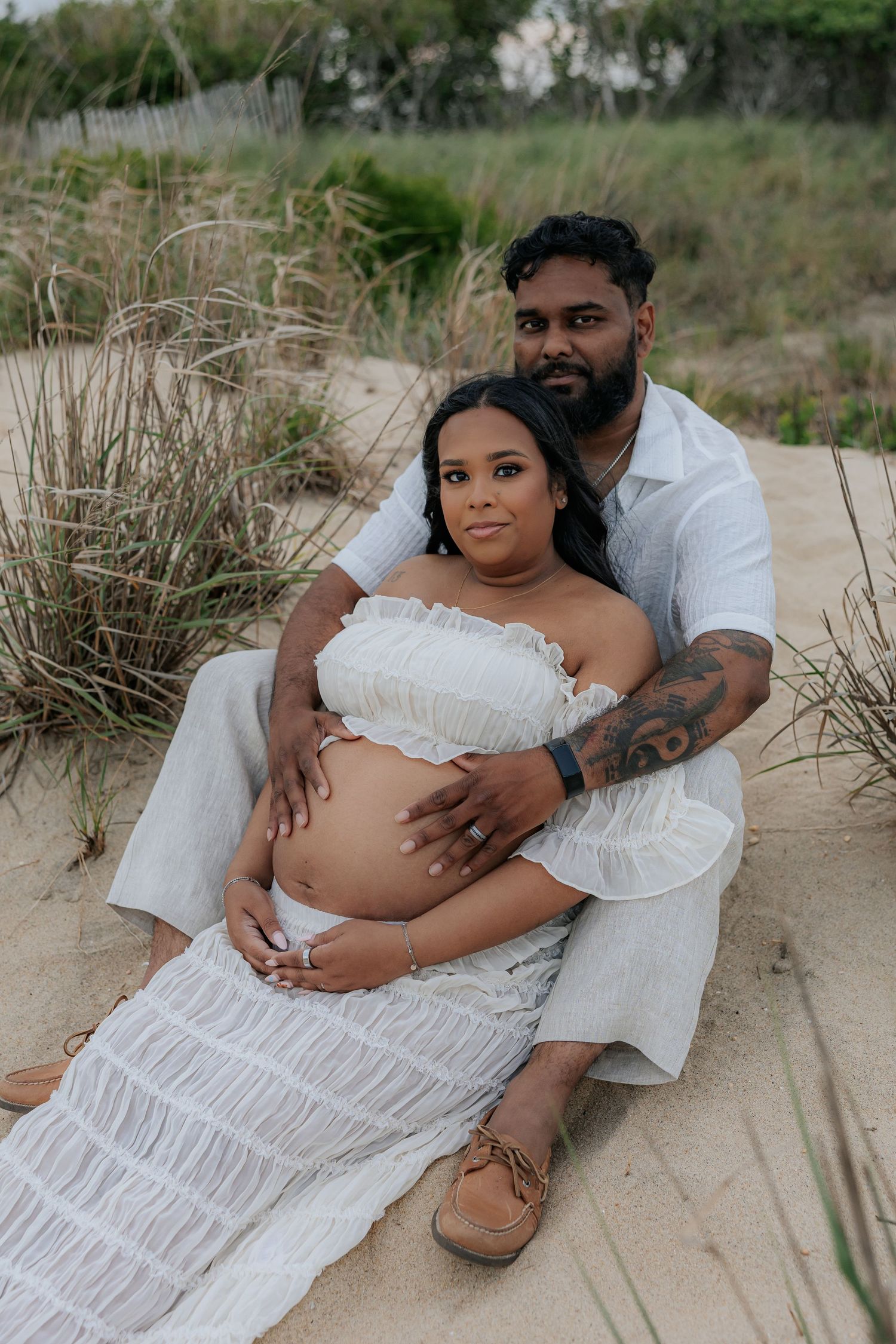 Couple sitting on sandy beach dunes, pregnant woman in white dress cradled by partner, maternity photo shoot outdoors.