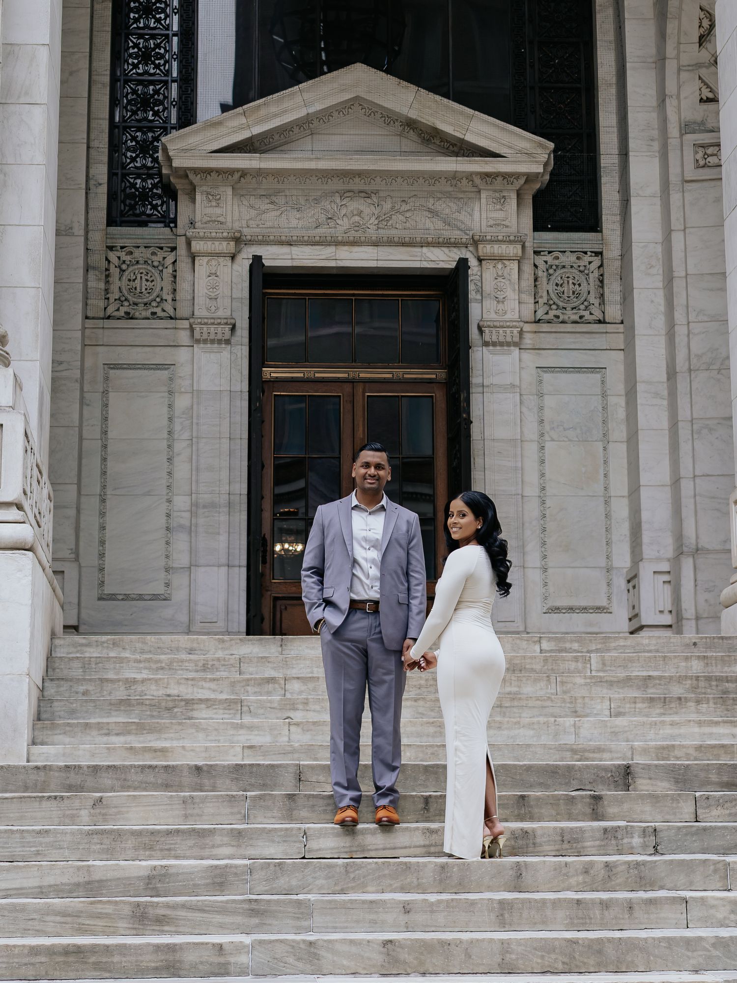 Elegant couple holding hands on the steps of a grand neoclassical stone building.