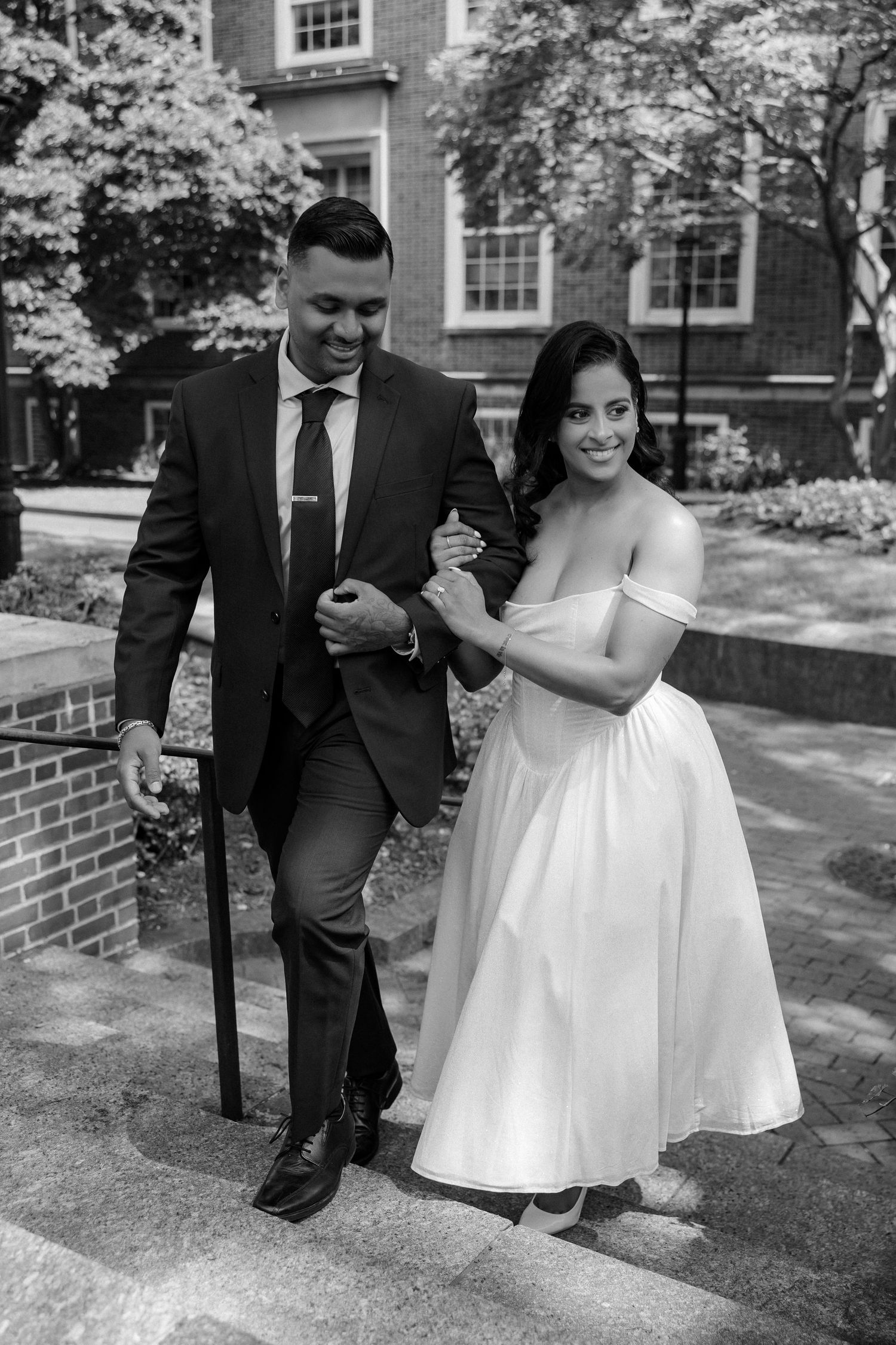 Black and white photo of a bride and groom walking down steps arm in arm outside a brick building.