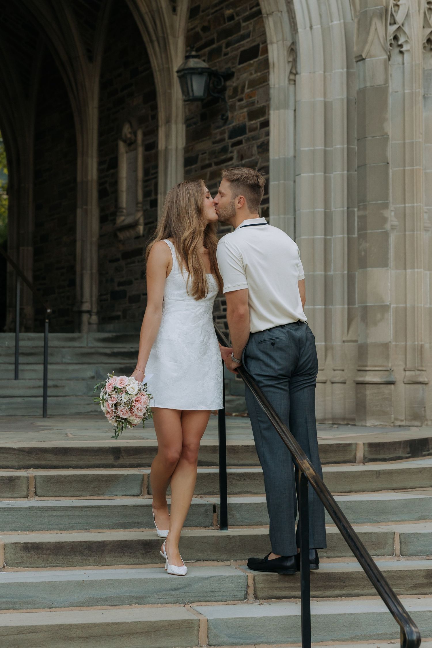 Bride and groom kissing on stone church steps, bride holding pink bouquet in white mini dress.
