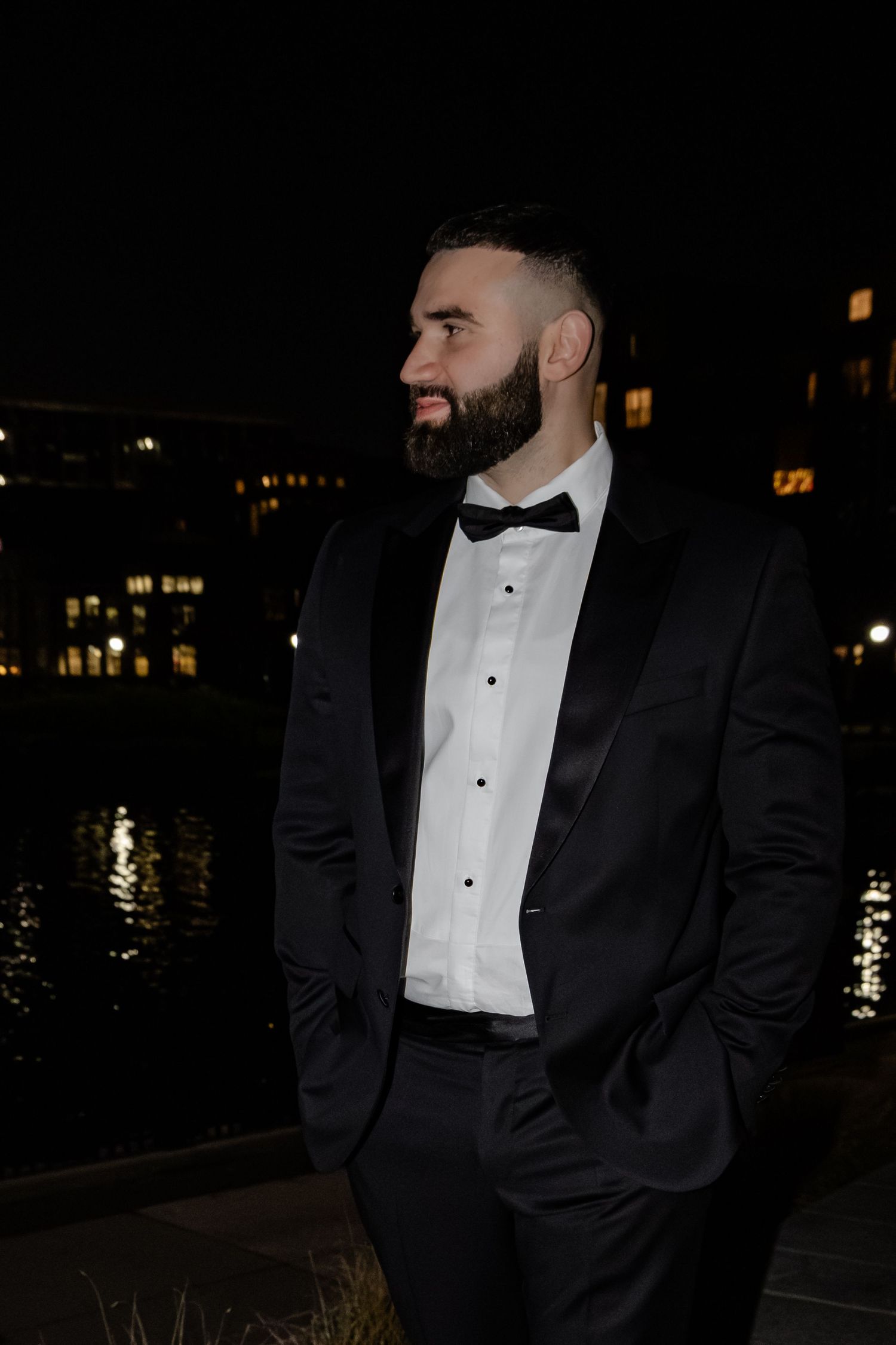Handsome groom in black tuxedo with bow tie standing outdoors near waterfront at night.