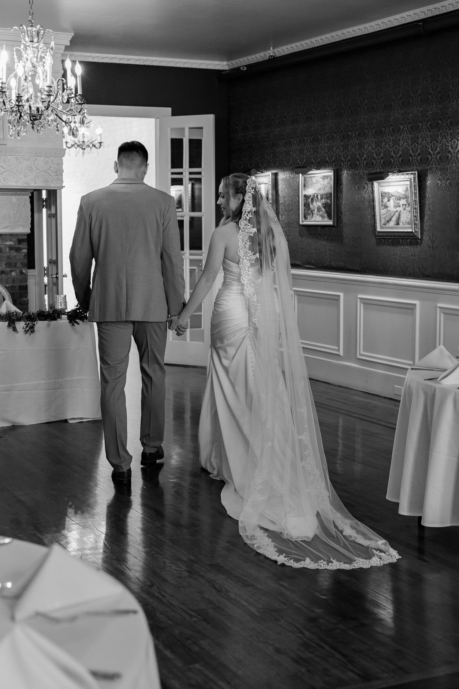 Bride and groom holding hands walking through elegant reception hall, black and white photo.