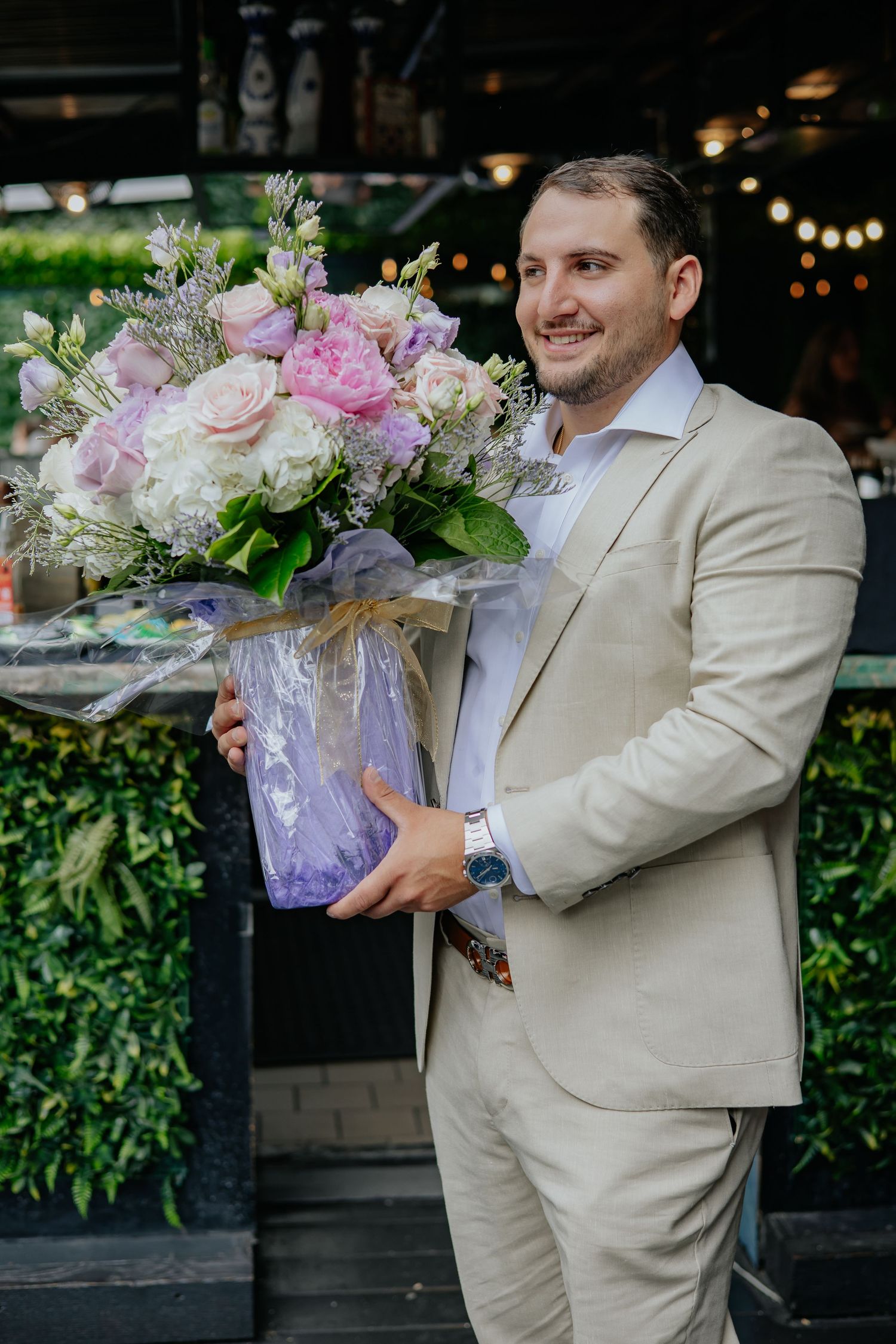 Smiling groom in beige suit holding a large pink and white floral arrangement in a vase.