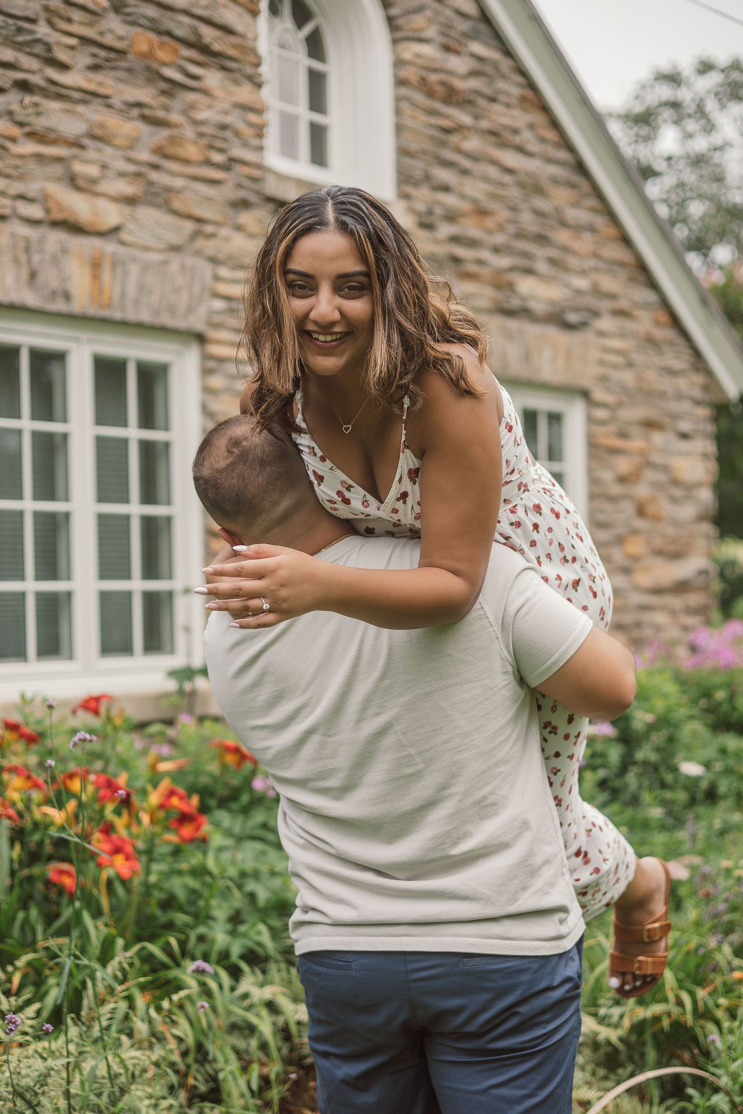 Man lifting smiling woman in floral dress outside stone building with garden flowers.