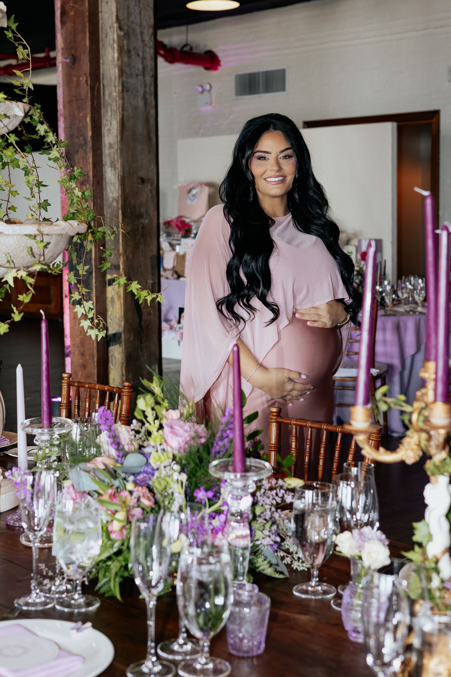Smiling pregnant woman in pink dress standing beside elegantly decorated baby shower table.