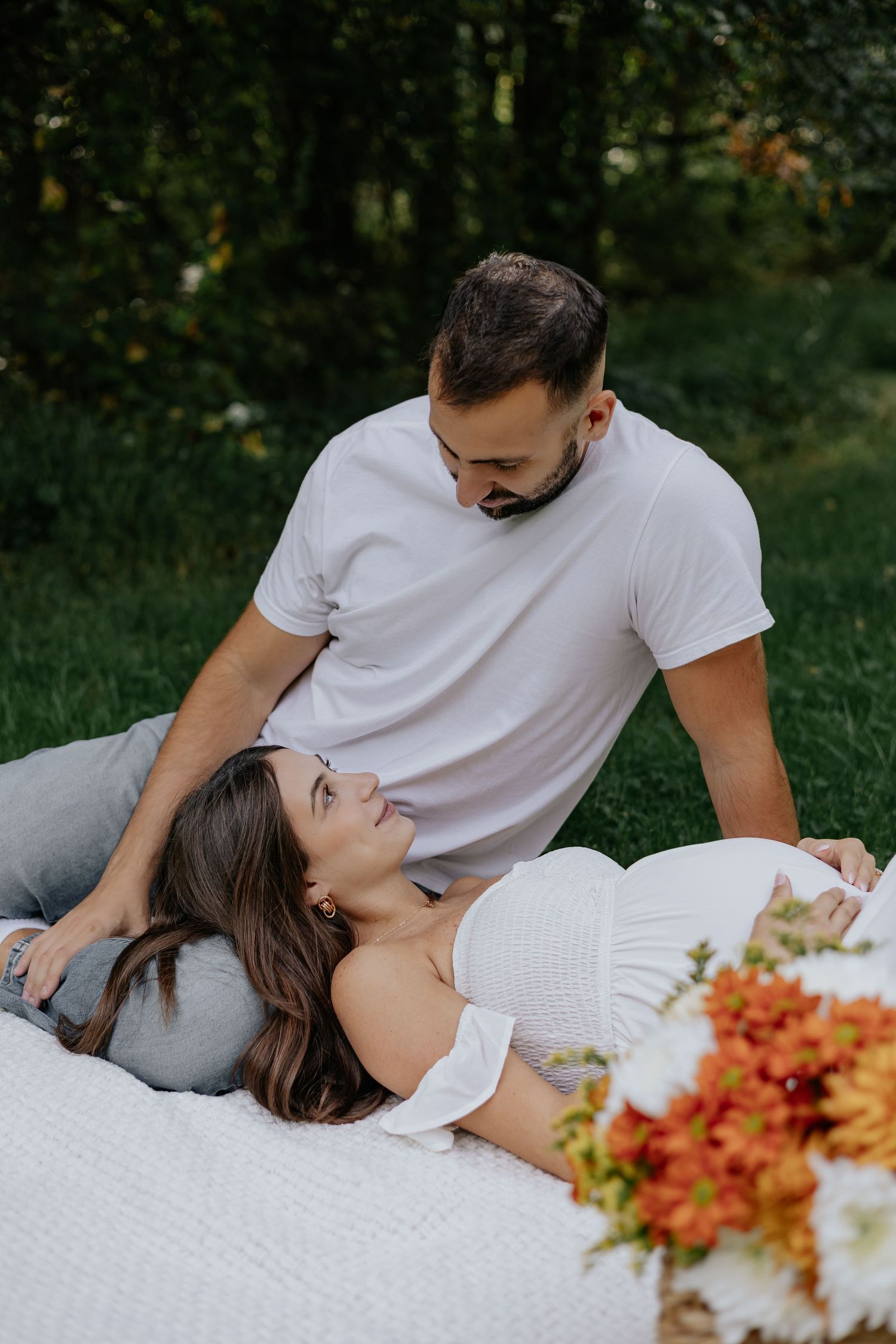 Pregnant woman lying on white blanket outdoors looking up at her partner surrounded by autumn flowers.