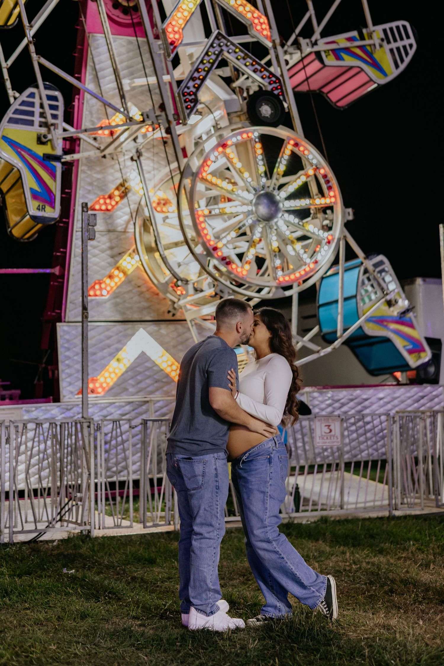 Pregnant couple sharing a kiss in front of illuminated carnival ride at night.