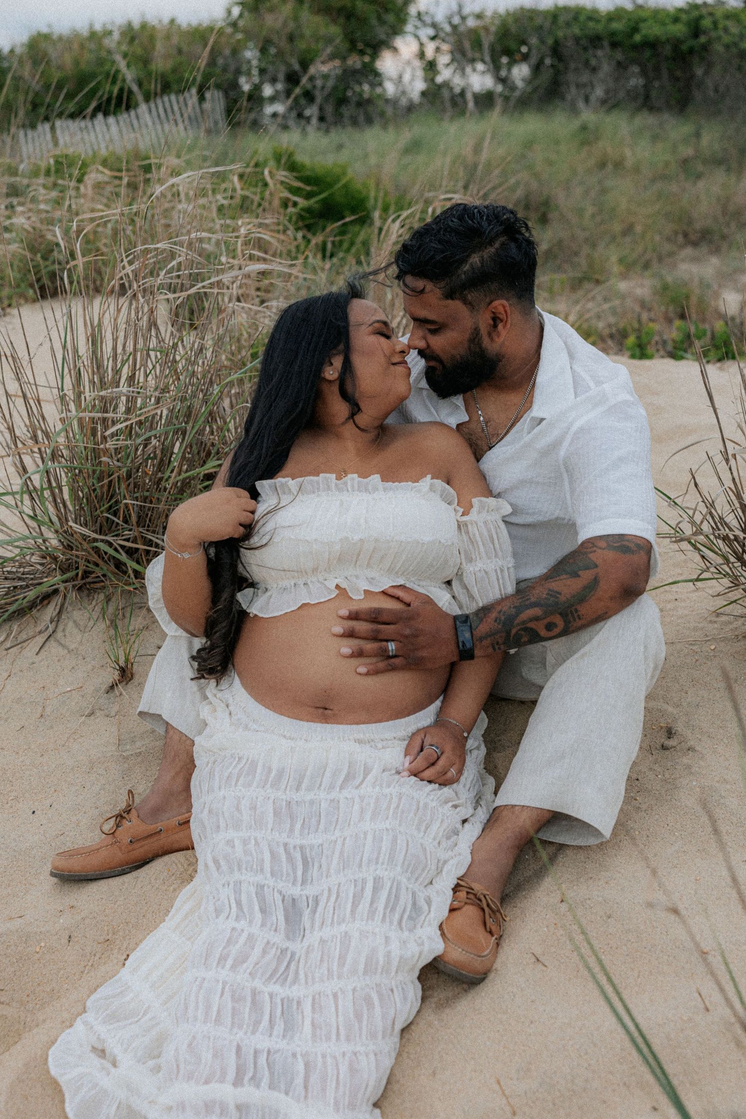 A couple in white outfits share a tender moment on sandy beach dunes, the woman visibly pregnant.