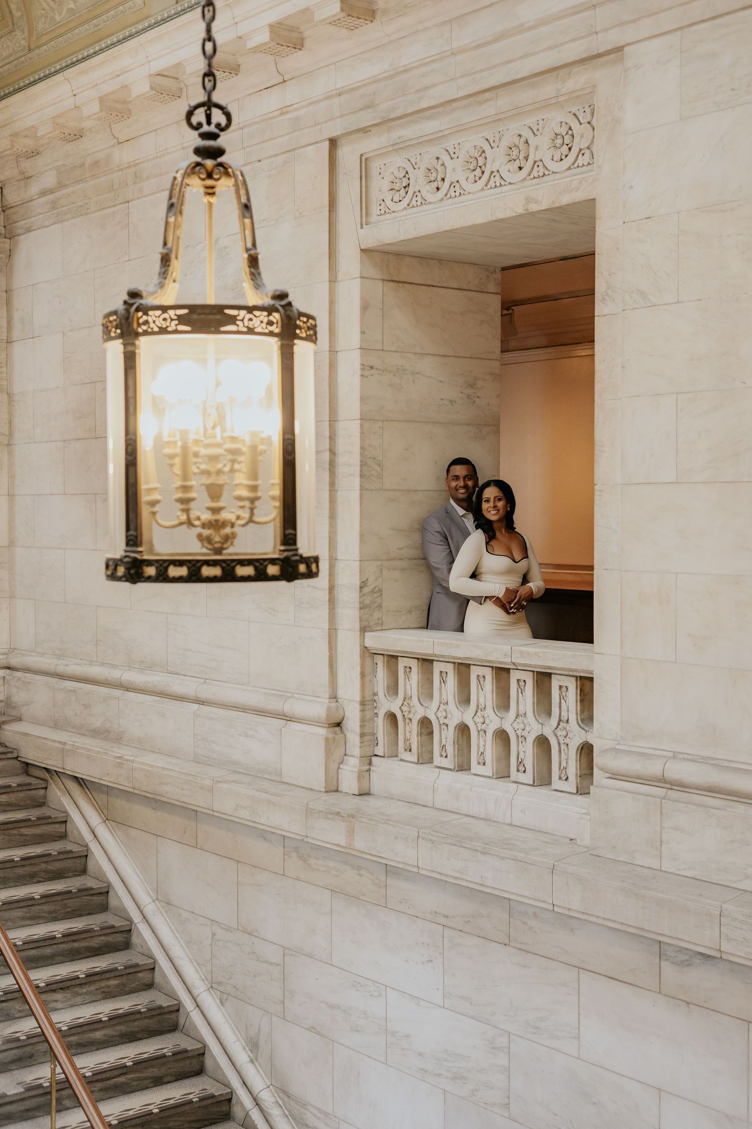 Couple posing in marble alcove balcony beside ornate gold lantern in grand classical building.