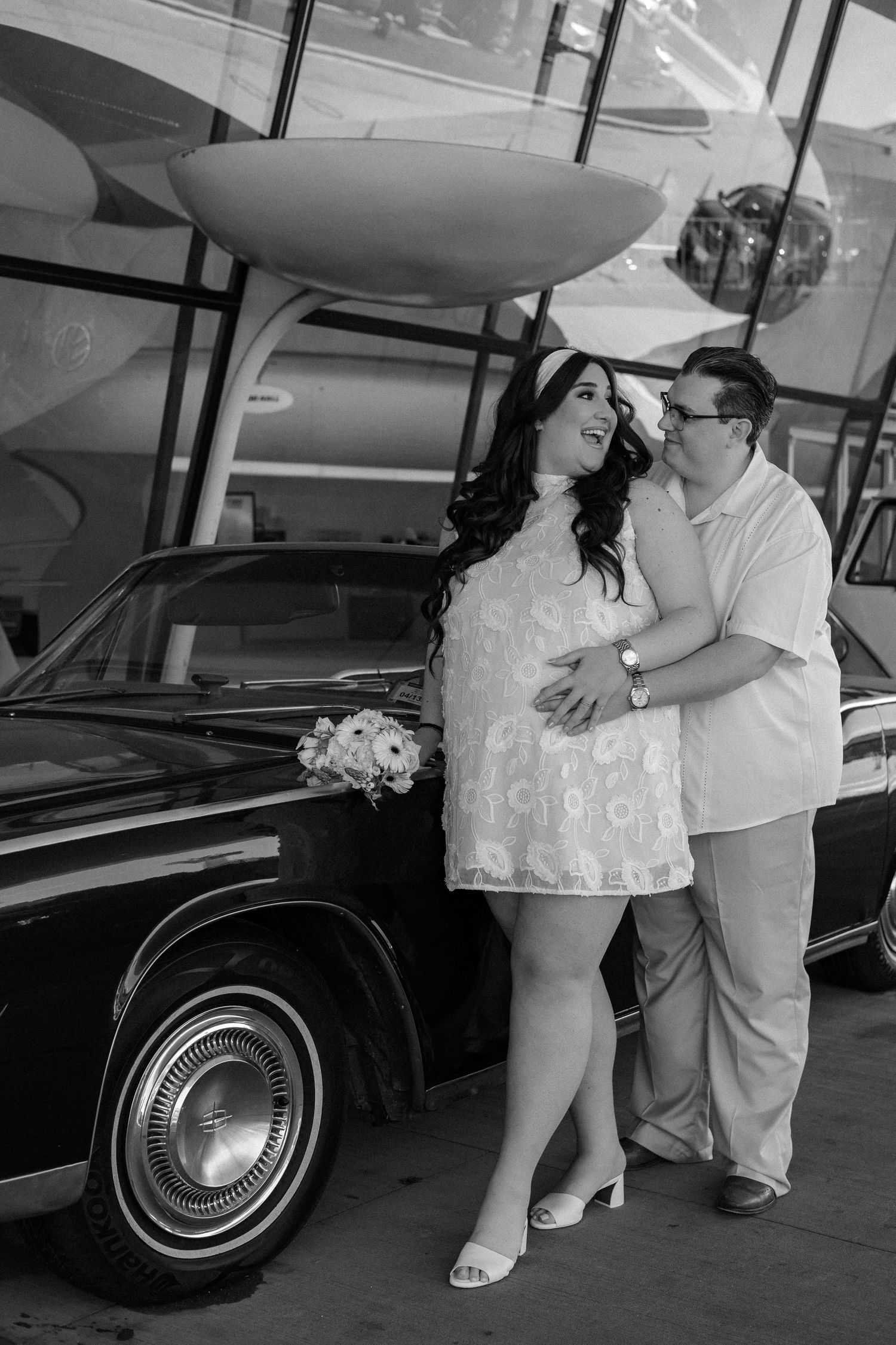 Black and white photo of couple embracing beside classic vintage car at retro airport terminal.