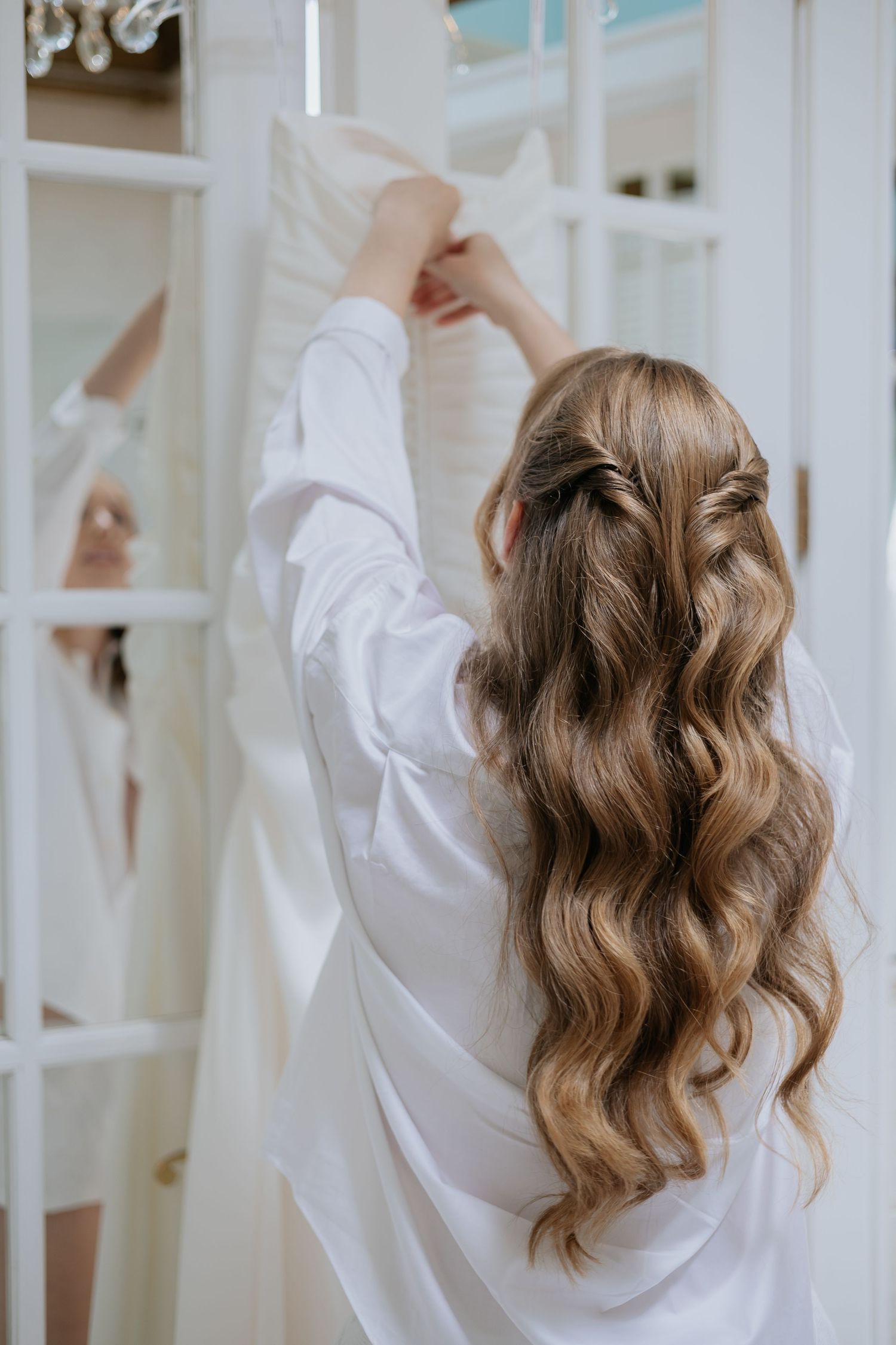 Bride in white robe reaching for wedding dress hanging on French door mirror.