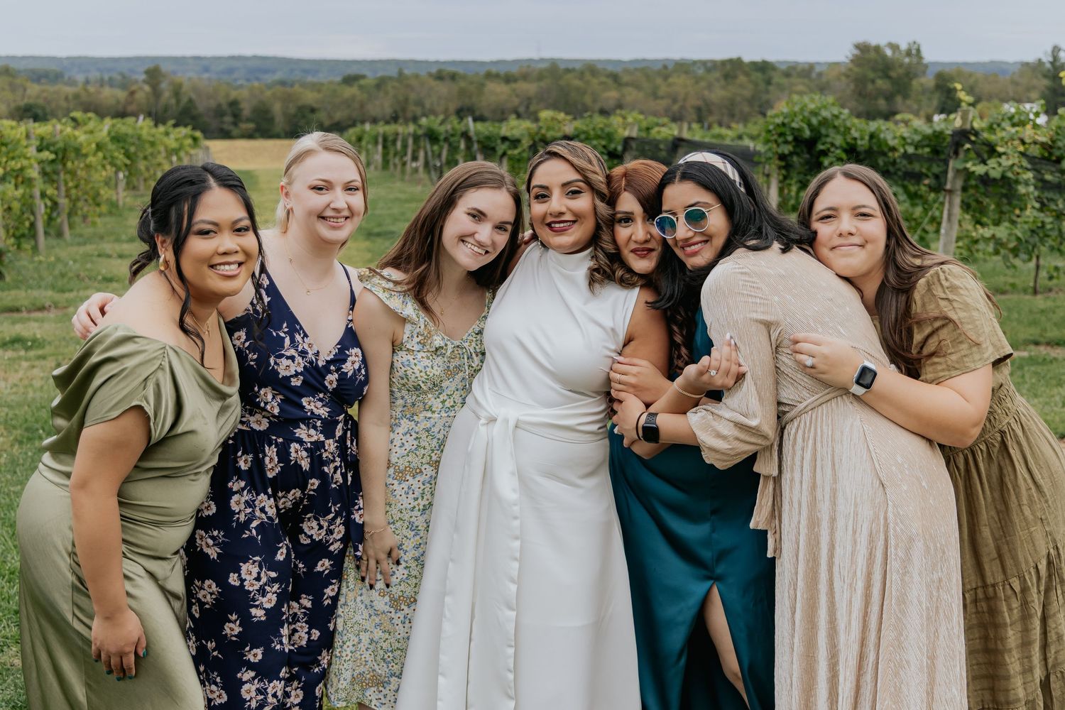 Seven smiling women in elegant dresses posing together at a sunny outdoor vineyard wedding.
