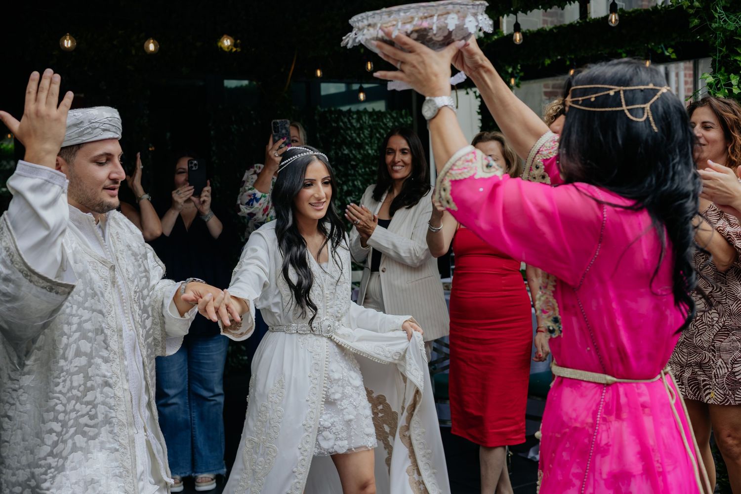Bride in white kaftan dancing joyfully with guests at traditional Moroccan wedding celebration.