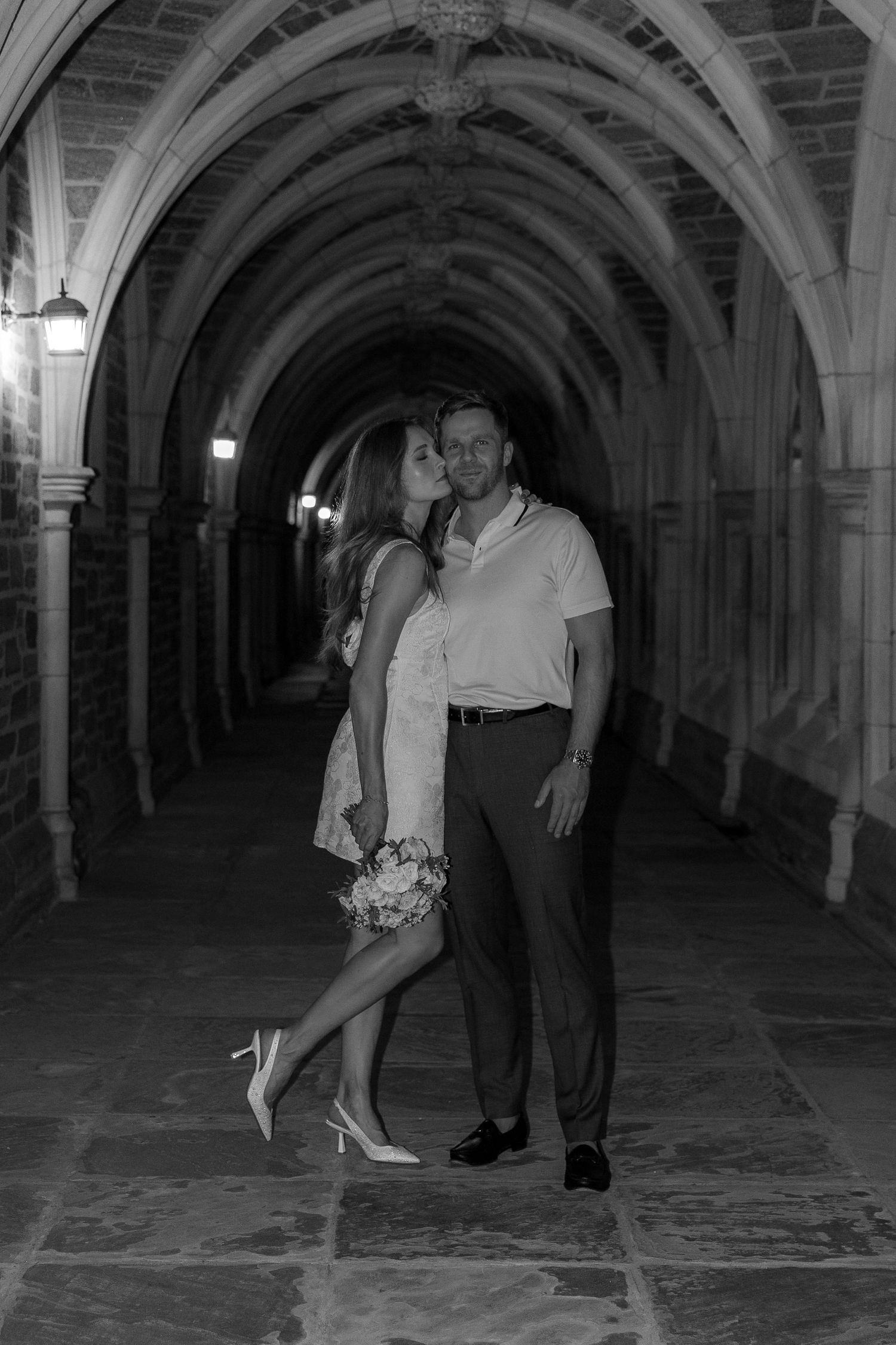 Black and white photo of a couple sharing a romantic kiss in an elegant stone archway corridor at night.