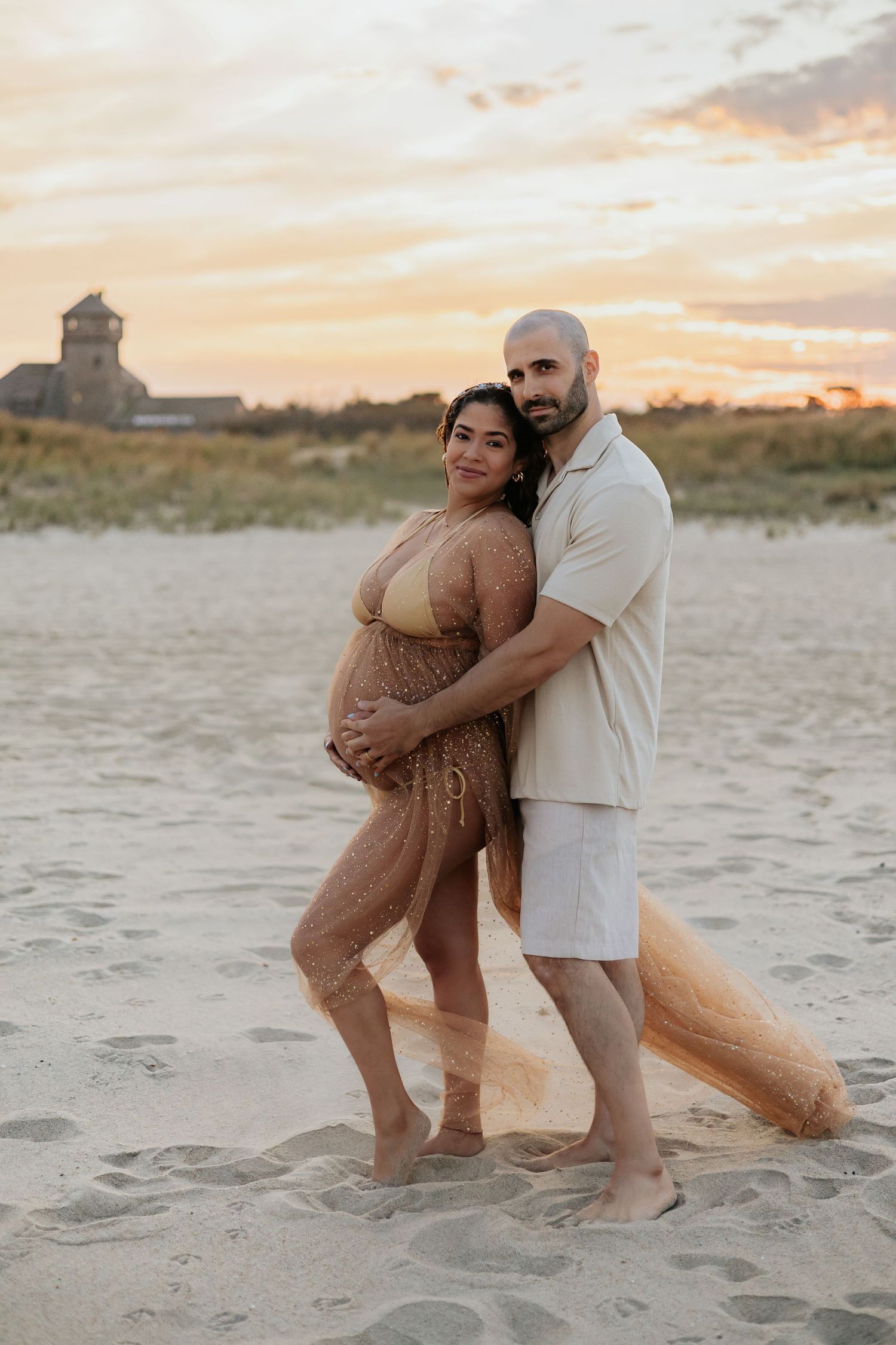 Expecting couple embracing at golden sunset on beach, woman wearing glittery gold maternity robe.