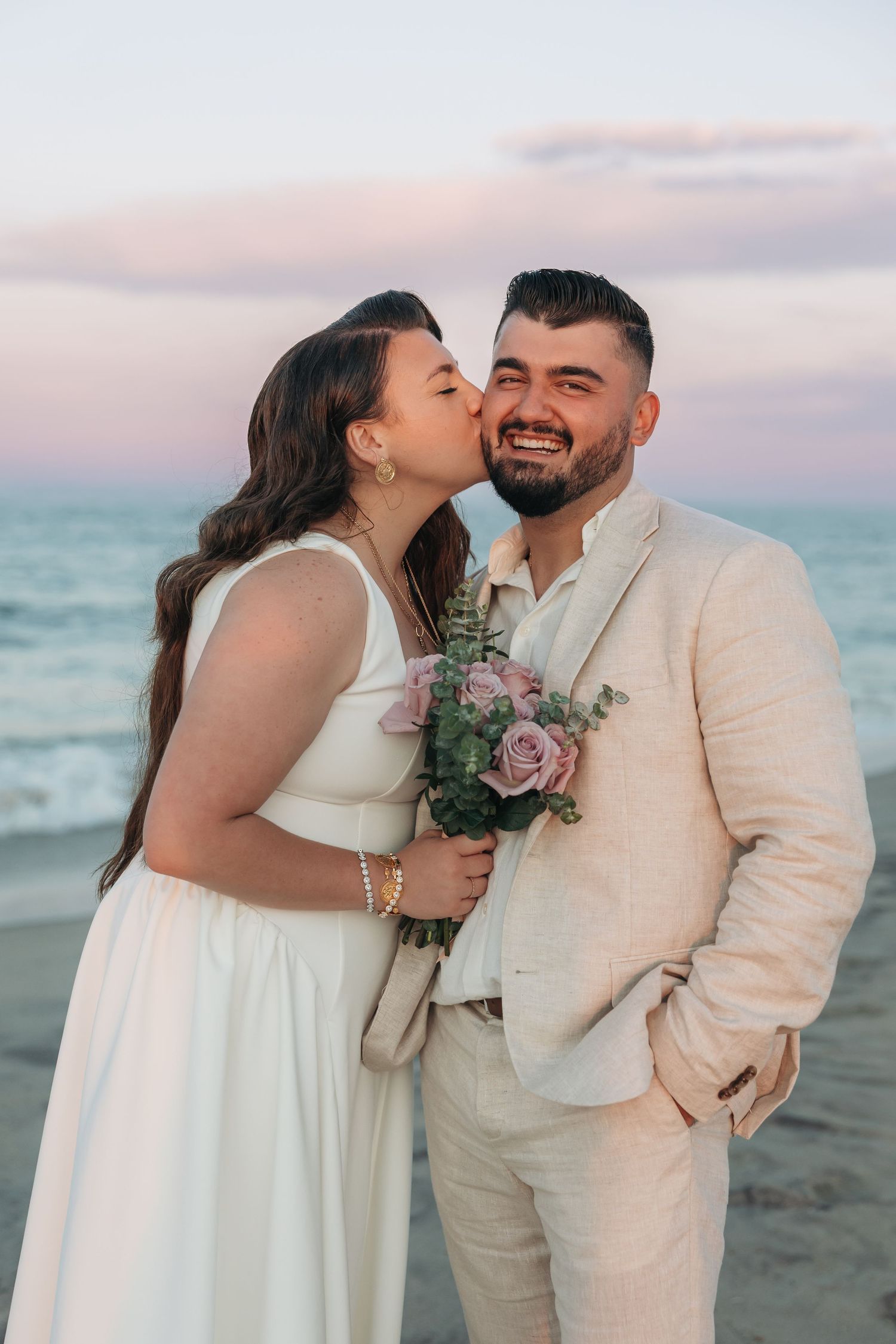Bride kissing groom on cheek at beach sunset, groom smiling holding pink rose bouquet, both in elegant wedding attire.