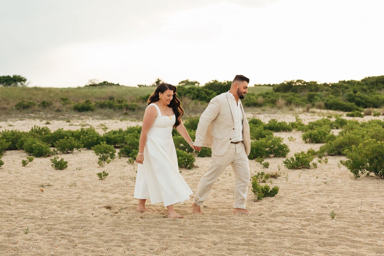Couple in wedding attire holding hands and walking barefoot on sandy beach dunes with green shrubs.