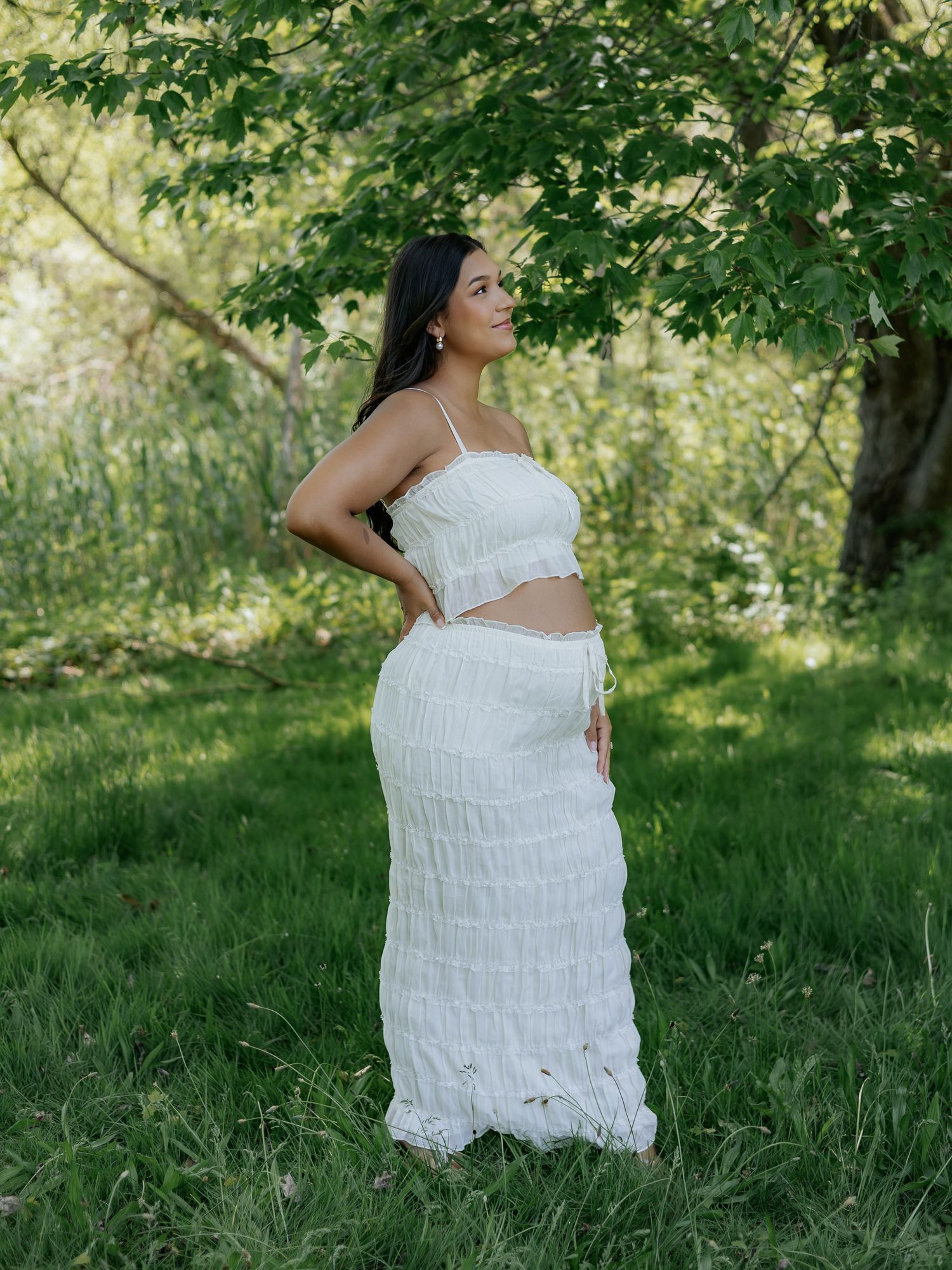 Woman in white crop top and maxi skirt posing outdoors in lush green garden setting.