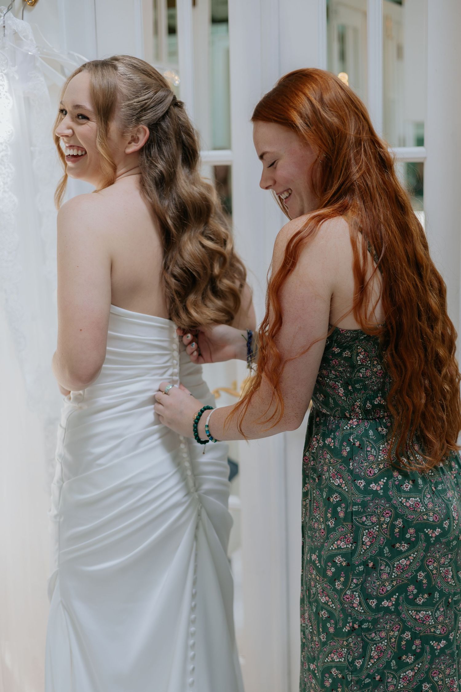 Bridesmaid in green floral dress laughing while buttoning bride's white wedding gown.
