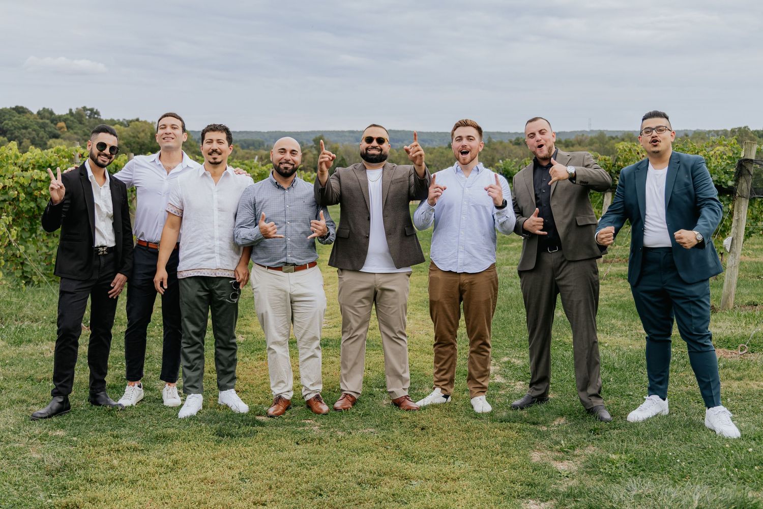 Seven men in semi-formal attire posing playfully at a lush green vineyard wedding celebration.