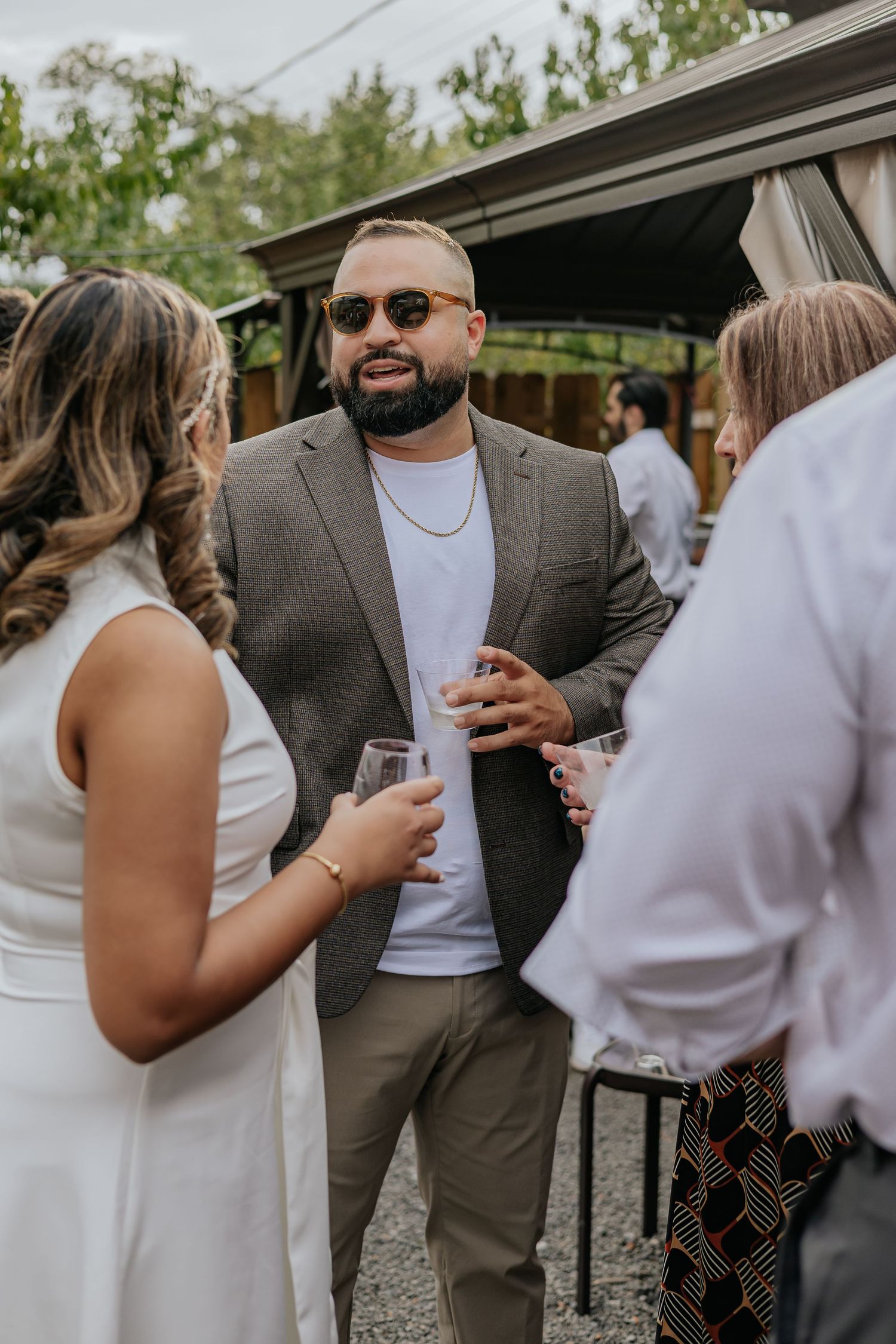 Bearded groom in gray blazer socializing with guests at outdoor engagement party.