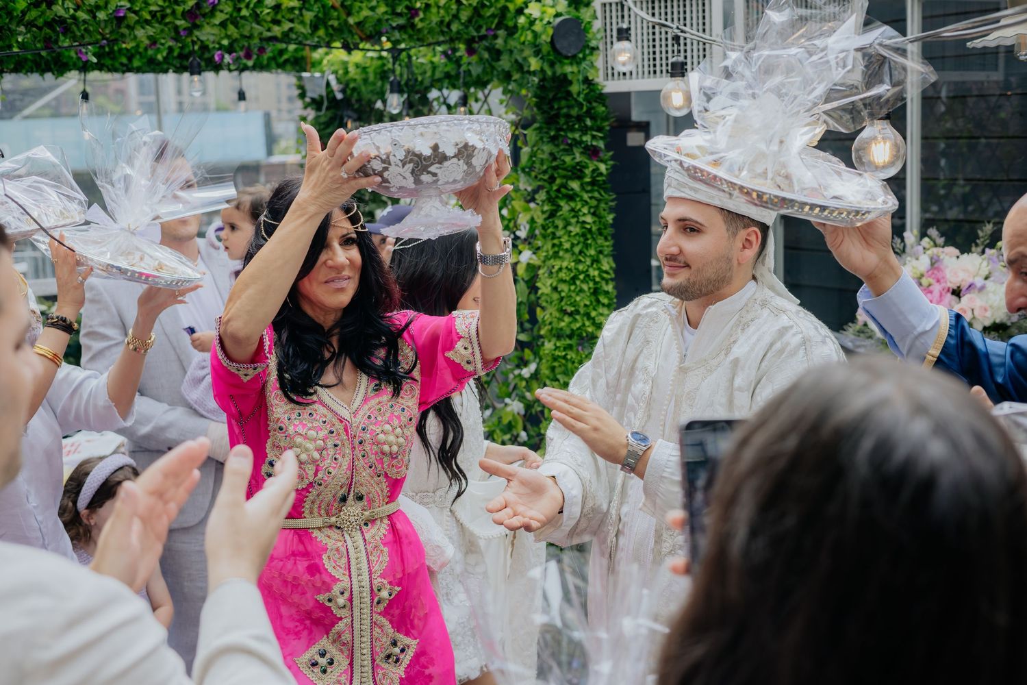 Bride in pink kaftan and groom in white traditional Moroccan attire celebrate with guests at a festive wedding ceremony.