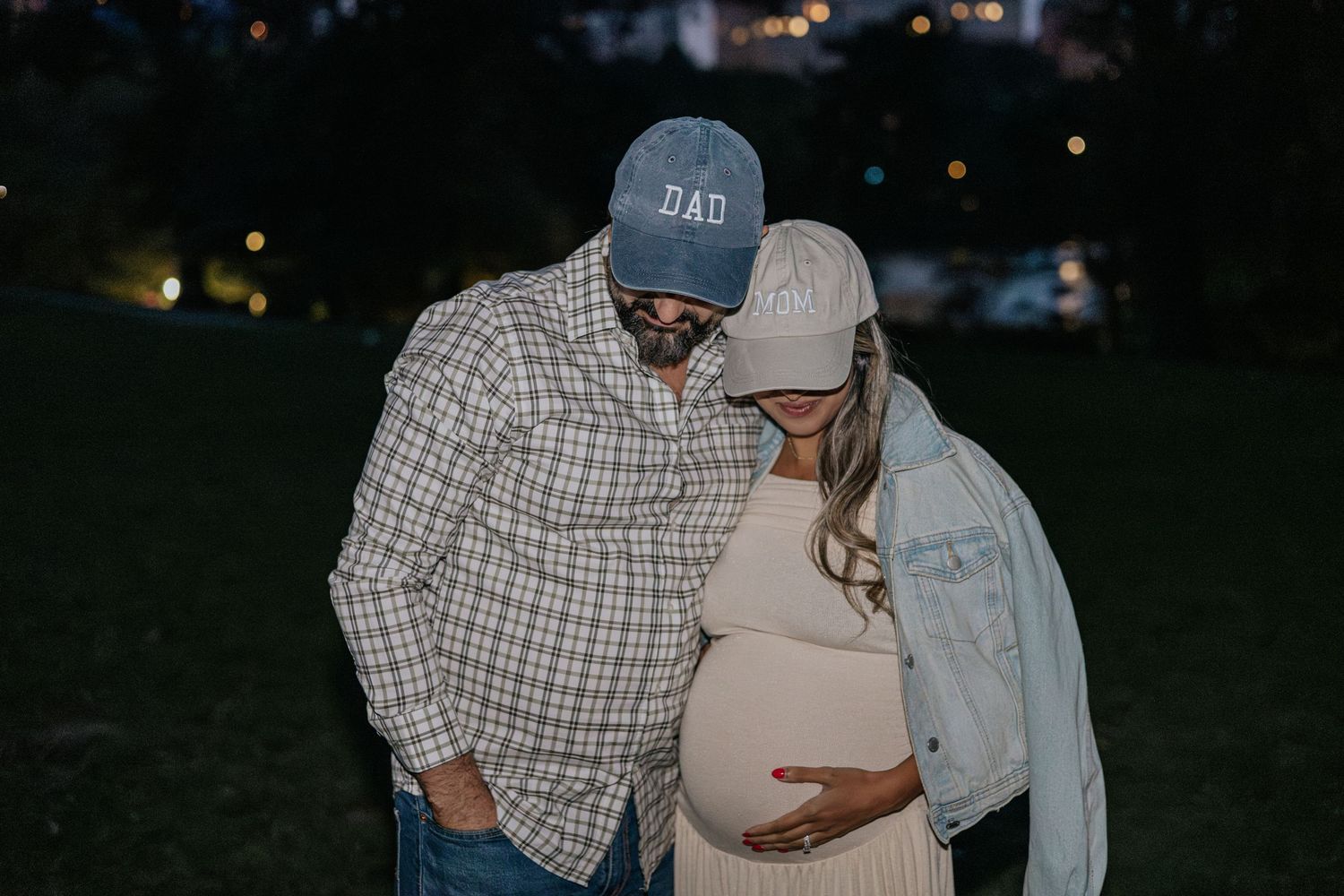 Couple in matching Dad and Mom caps looking down at pregnant belly at night.
