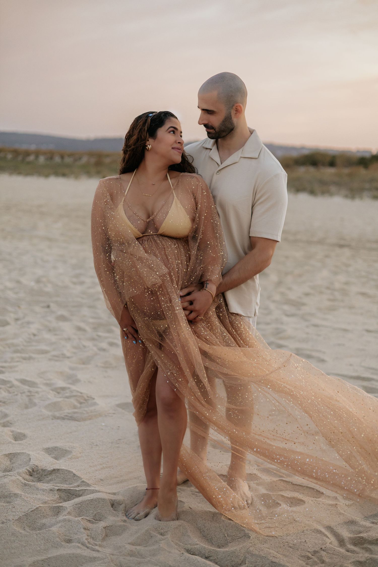 Pregnant couple embracing on sandy beach at sunset, woman in gold glitter tulle maternity gown.
