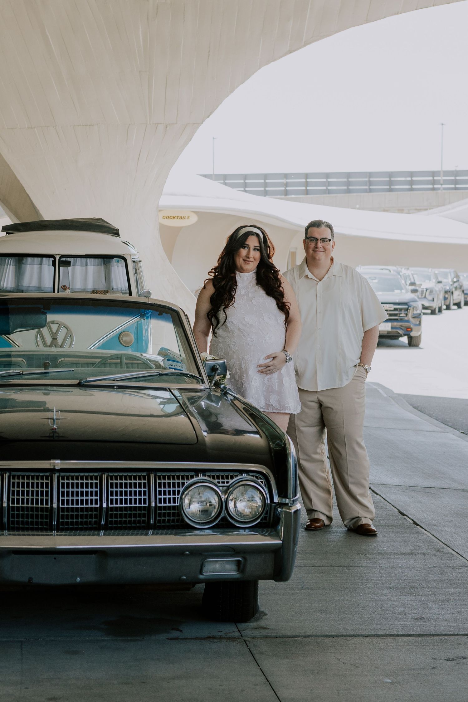 Smiling pregnant couple stands beside classic black vintage car at retro TWA Hotel terminal.