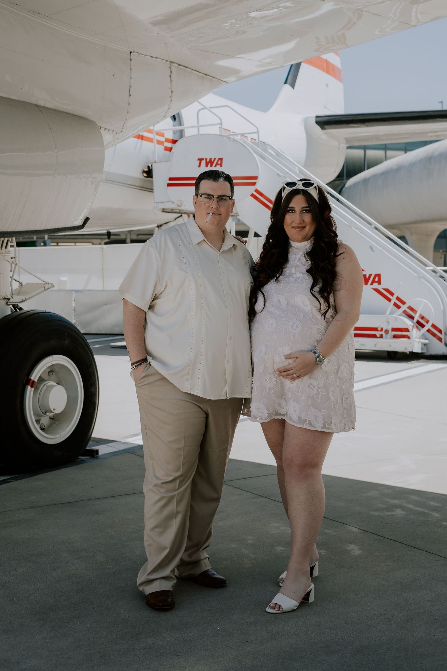 Couple posing together in front of a vintage TWA airplane on a sunny day at an airport.