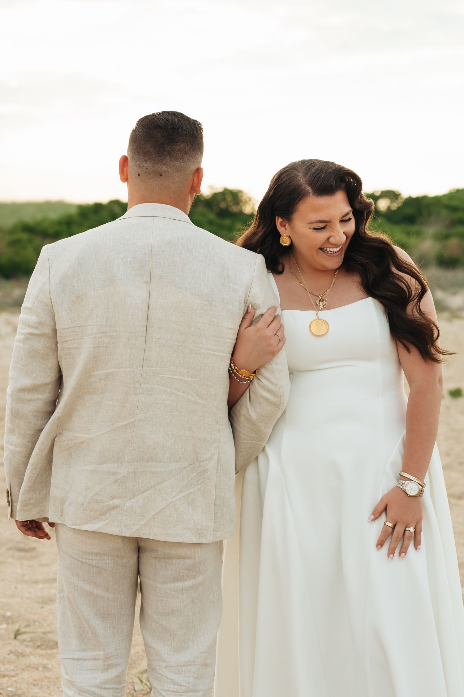Bride in white gown smiles at groom in beige linen suit during outdoor wedding photo session.