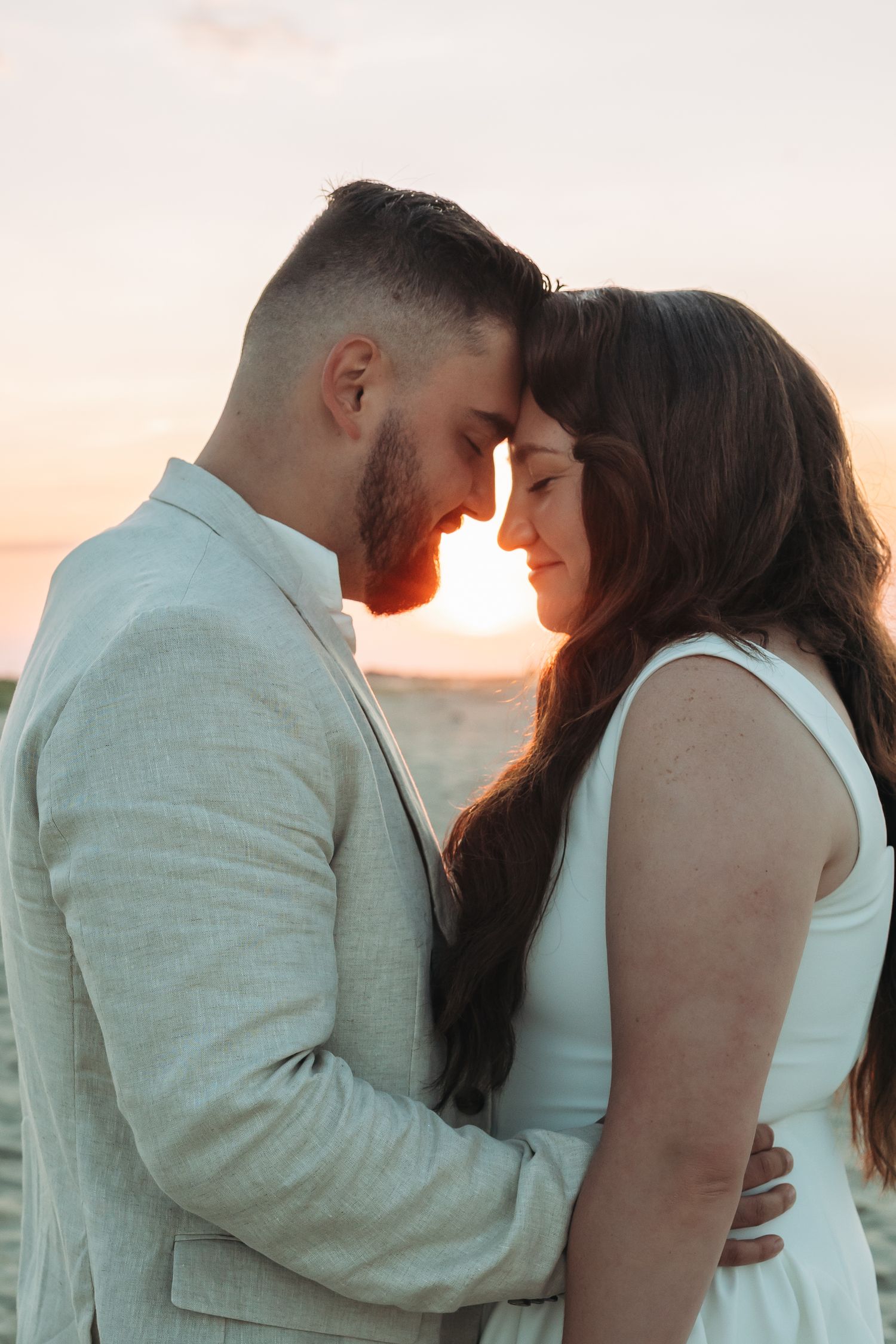 A romantic couple in wedding attire touching foreheads at sunset on a beach, sharing an intimate moment.