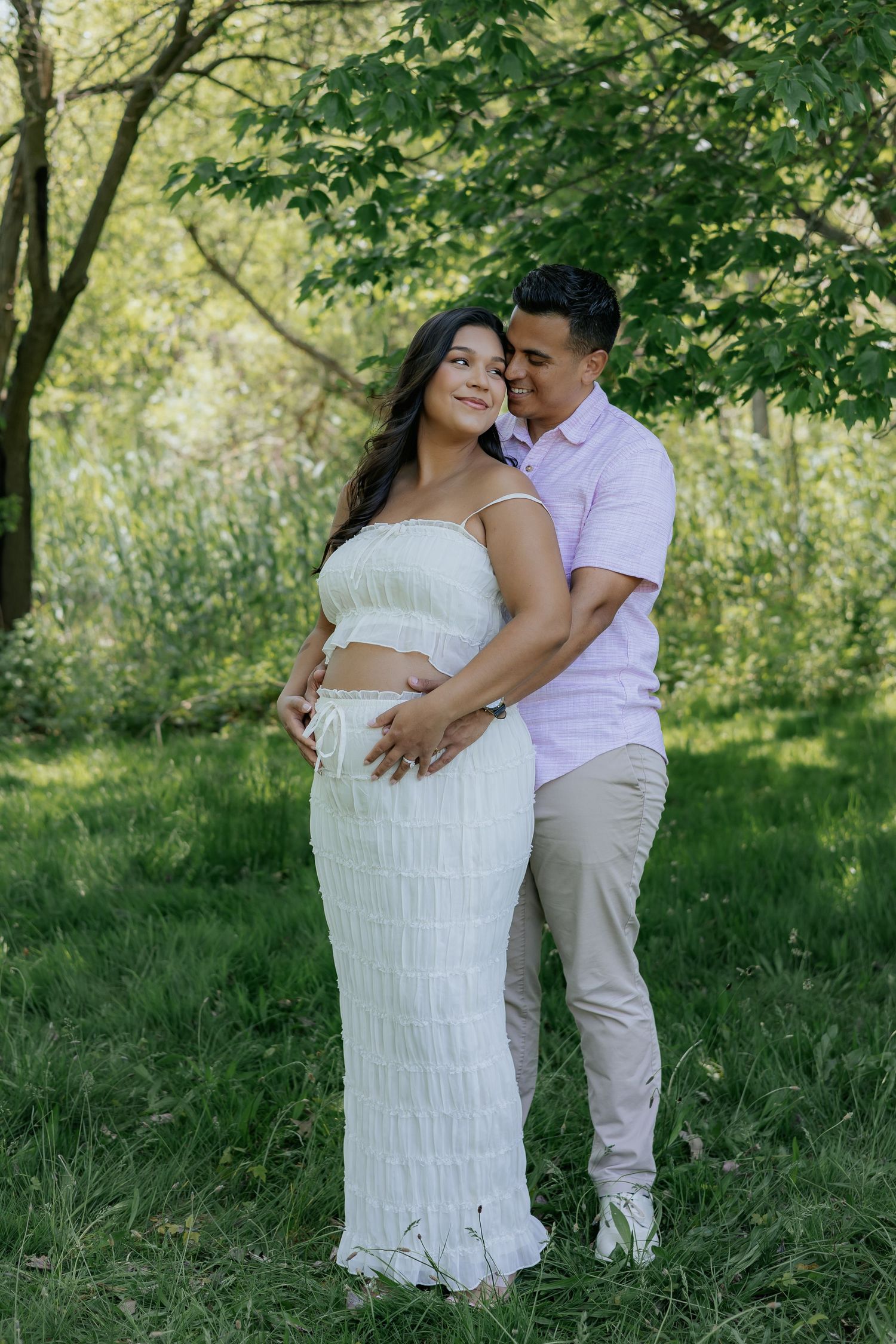 Couple poses in lush green park, pregnant woman in white dress, partner embracing her from behind.