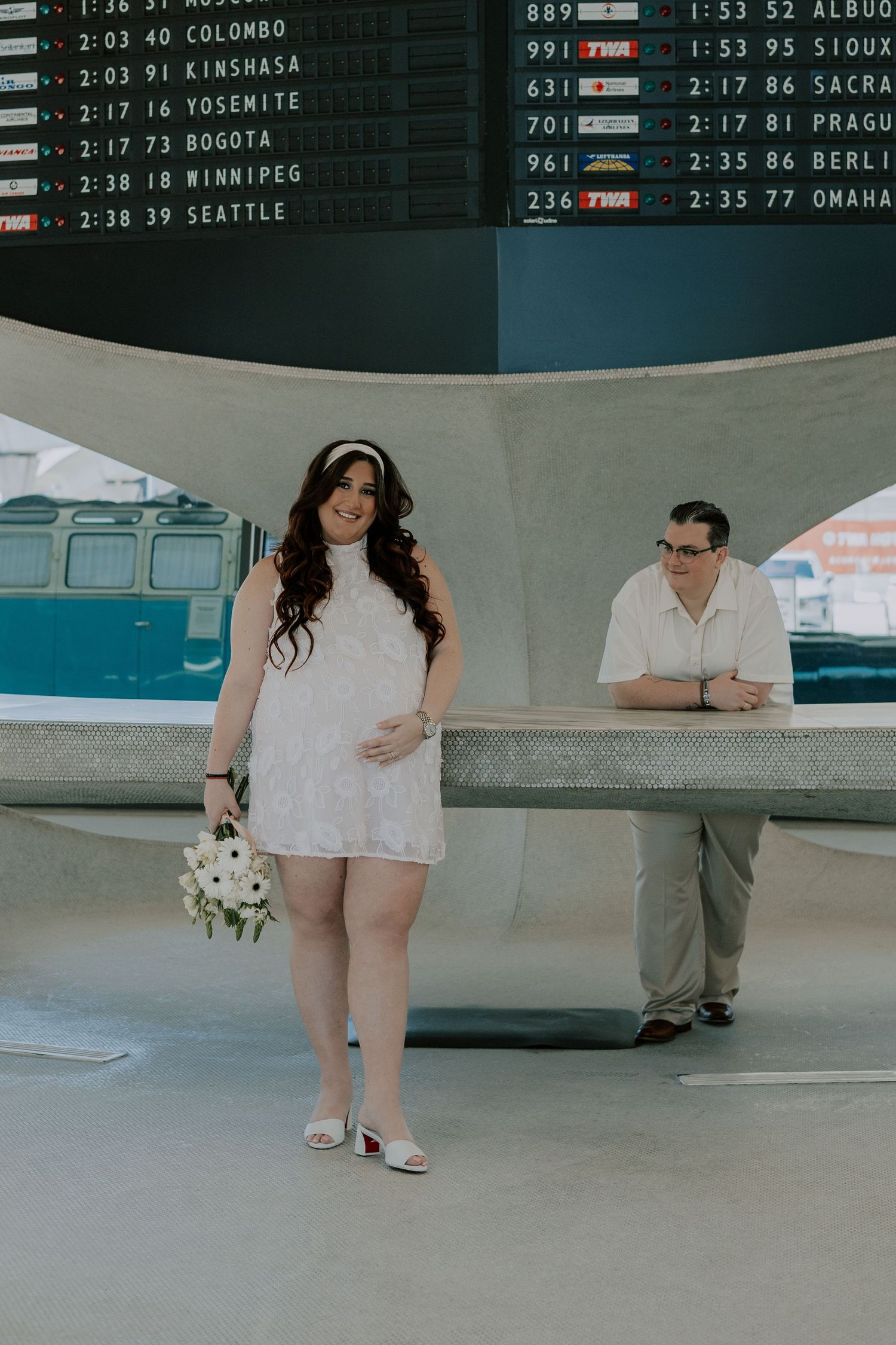 Pregnant bride in white mini dress holds bouquet near vintage airport flight departures board.