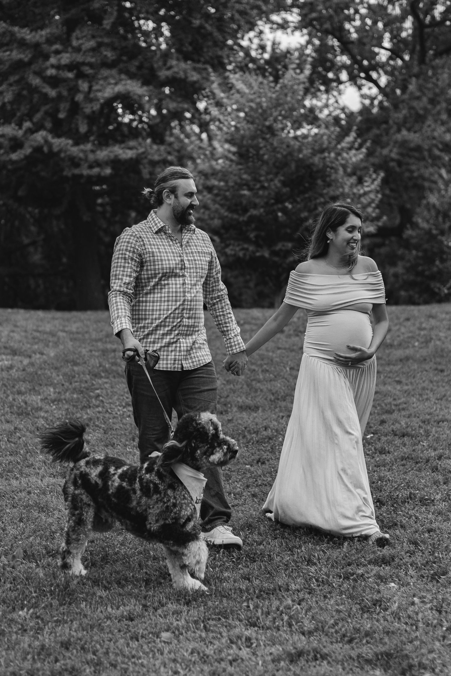Black and white photo of pregnant couple walking their dog in a lush park.