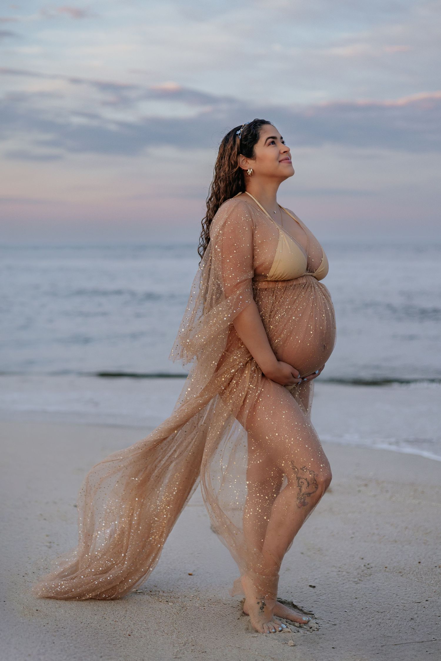 Pregnant woman in gold sparkly sheer robe standing on beach at sunset holding baby bump gracefully.