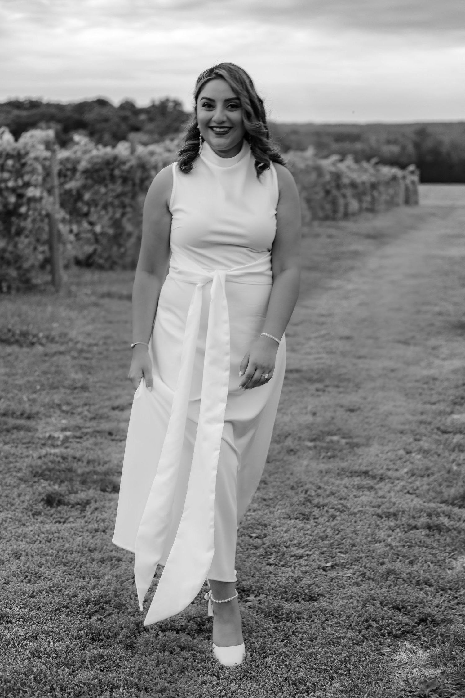 Black and white portrait of smiling woman in white sleeveless dress walking through vineyard path.