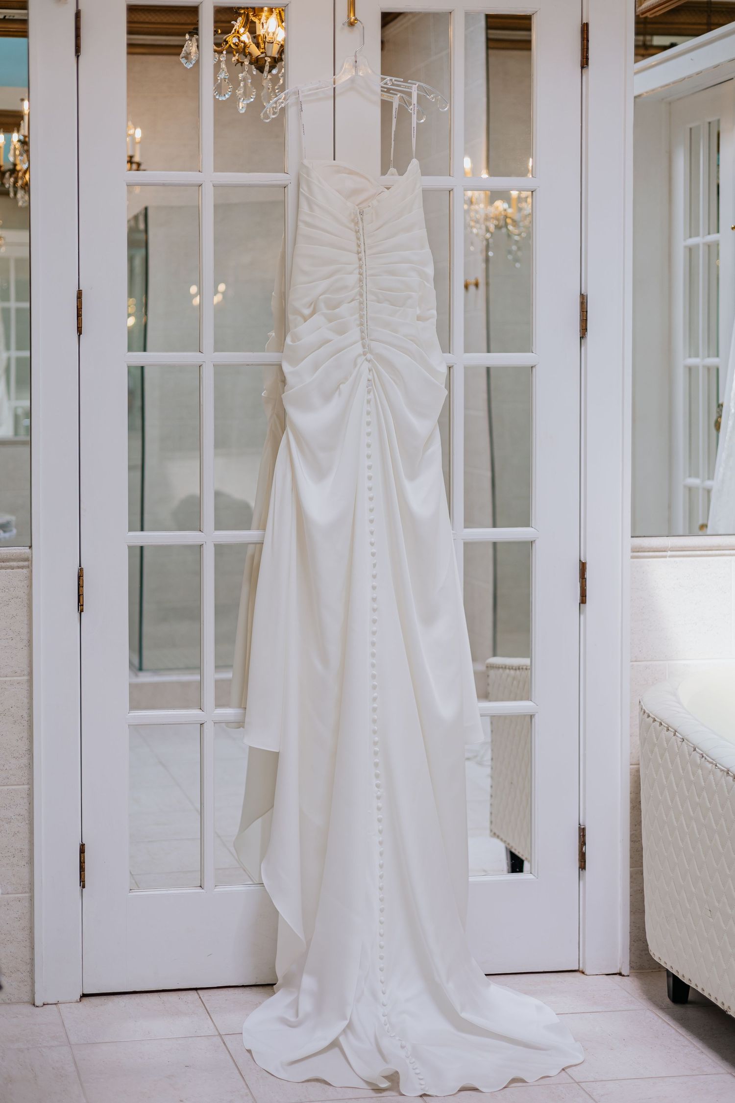 Elegant white satin wedding gown with button back hanging on French doors.