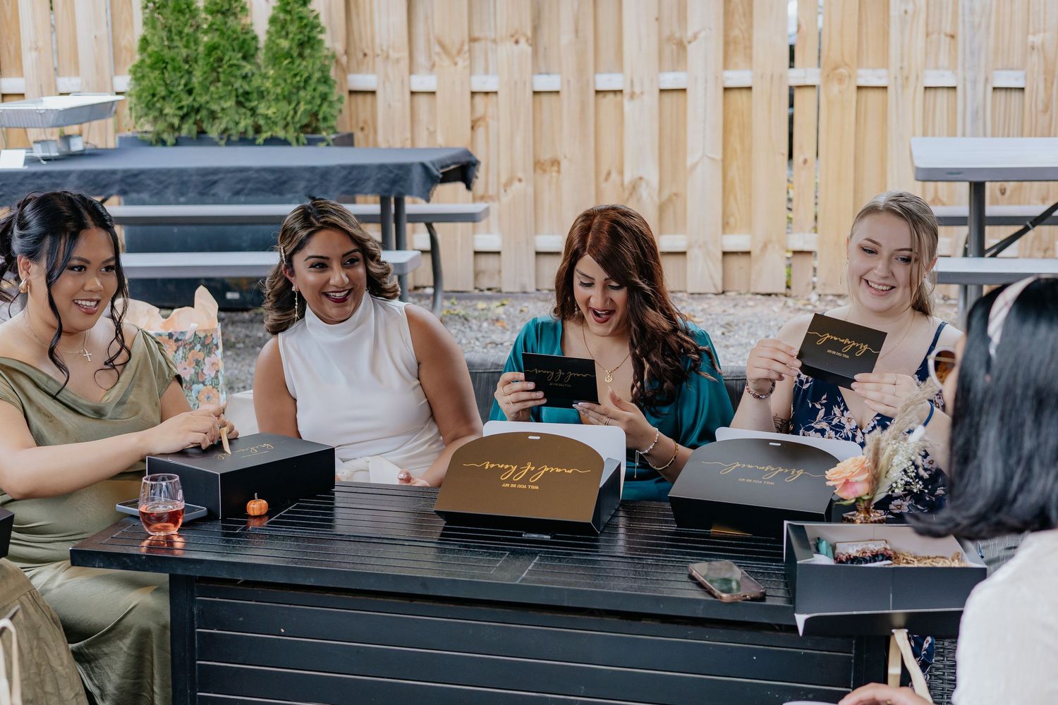 Group of bridesmaids opening black and gold bridesmaid proposal boxes at outdoor table.