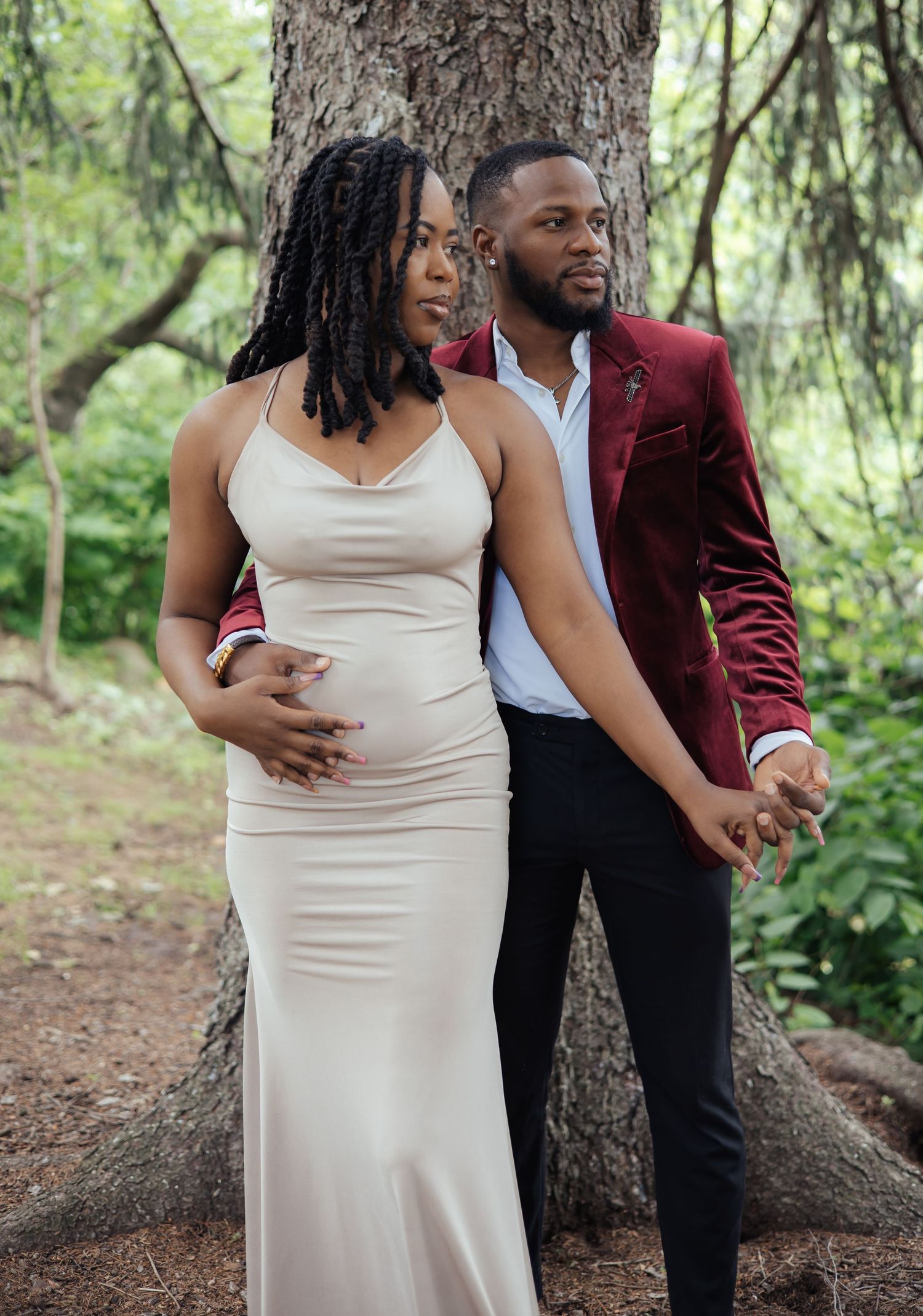 Pregnant woman in a champagne gown and her partner in a burgundy blazer posing in a forest.