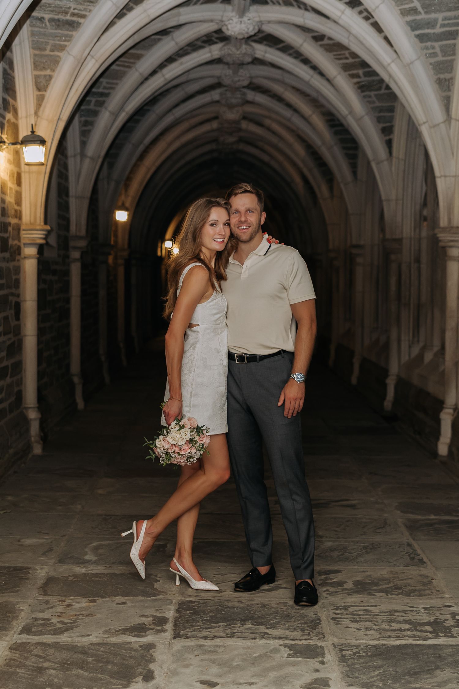 Couple posing under stone Gothic arches at night, woman in white dress holding flowers, man in beige polo and gray pants.