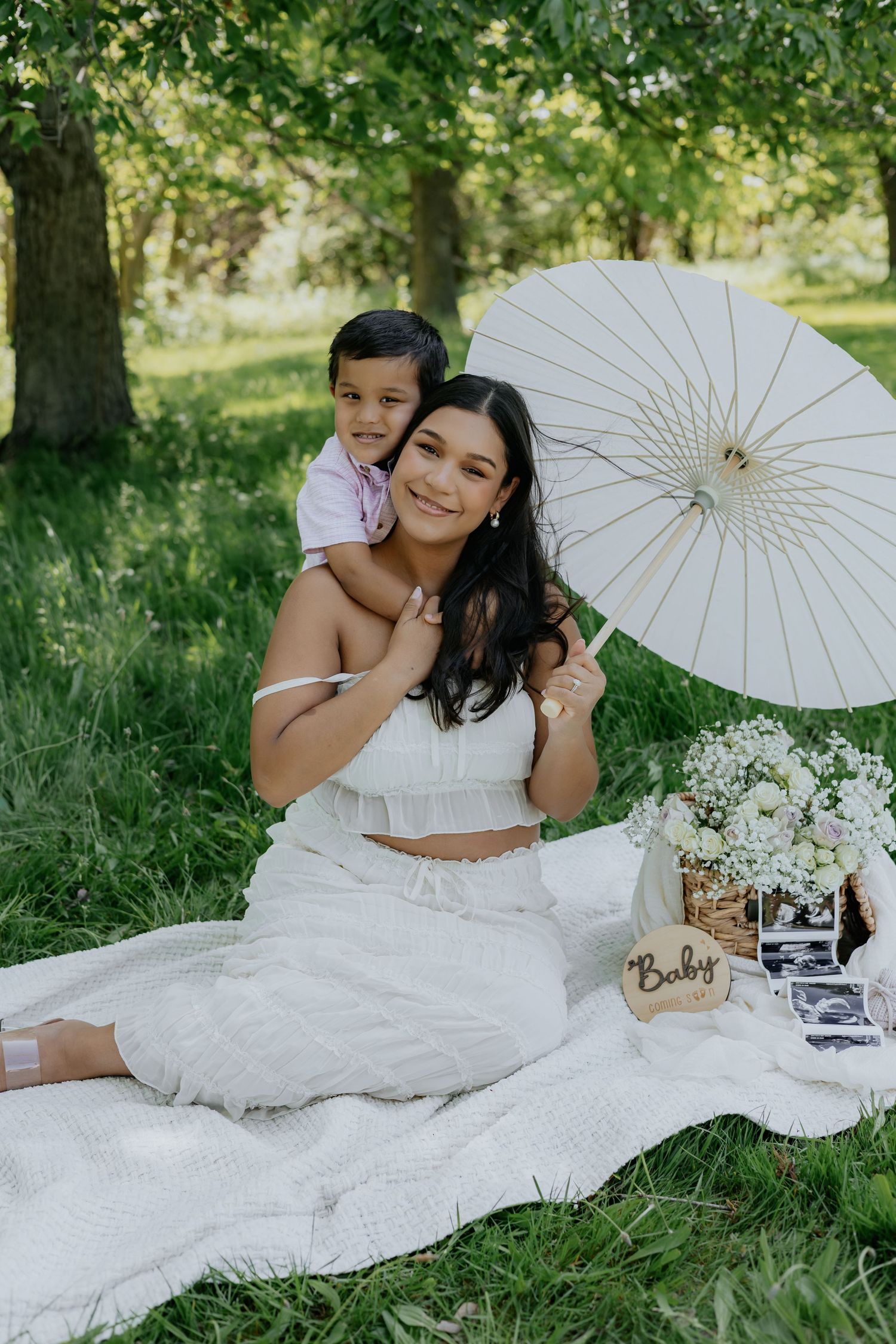Pregnant woman in white outfit holds parasol at outdoor picnic with toddler, baby announcement sign and ultrasound photos nearby.