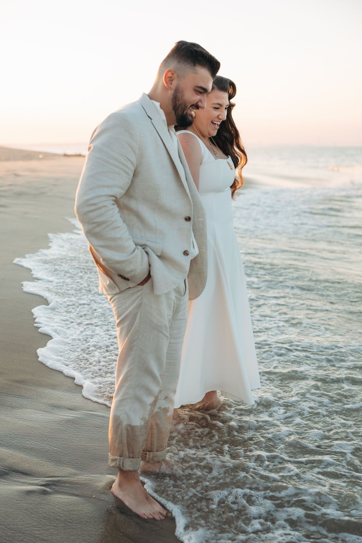 Couple in wedding attire standing barefoot on a sandy beach at sunset, waves gently washing over their feet.